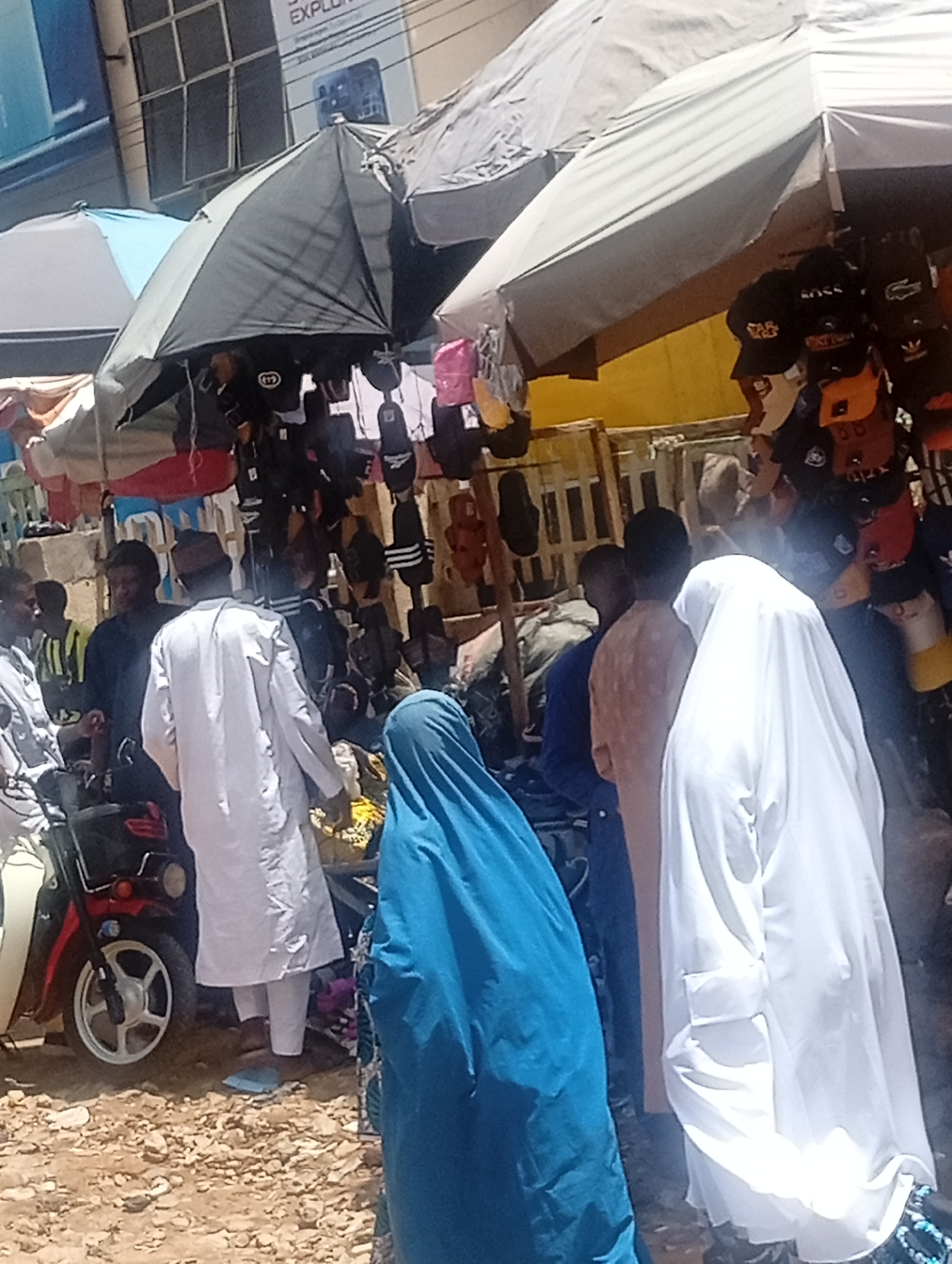 The image shows a busy street scene in Maiburiji, Nigeria. Several people are visible, including two women wearing hijabs (one blue and one white). Men are also present, some wearing traditional clothing. A red motorcycle is parked on the side. Overhead, several umbrellas provide shade over what appears to be a market stall selling items like hats and bags. The ground is uneven and possibly unpaved.