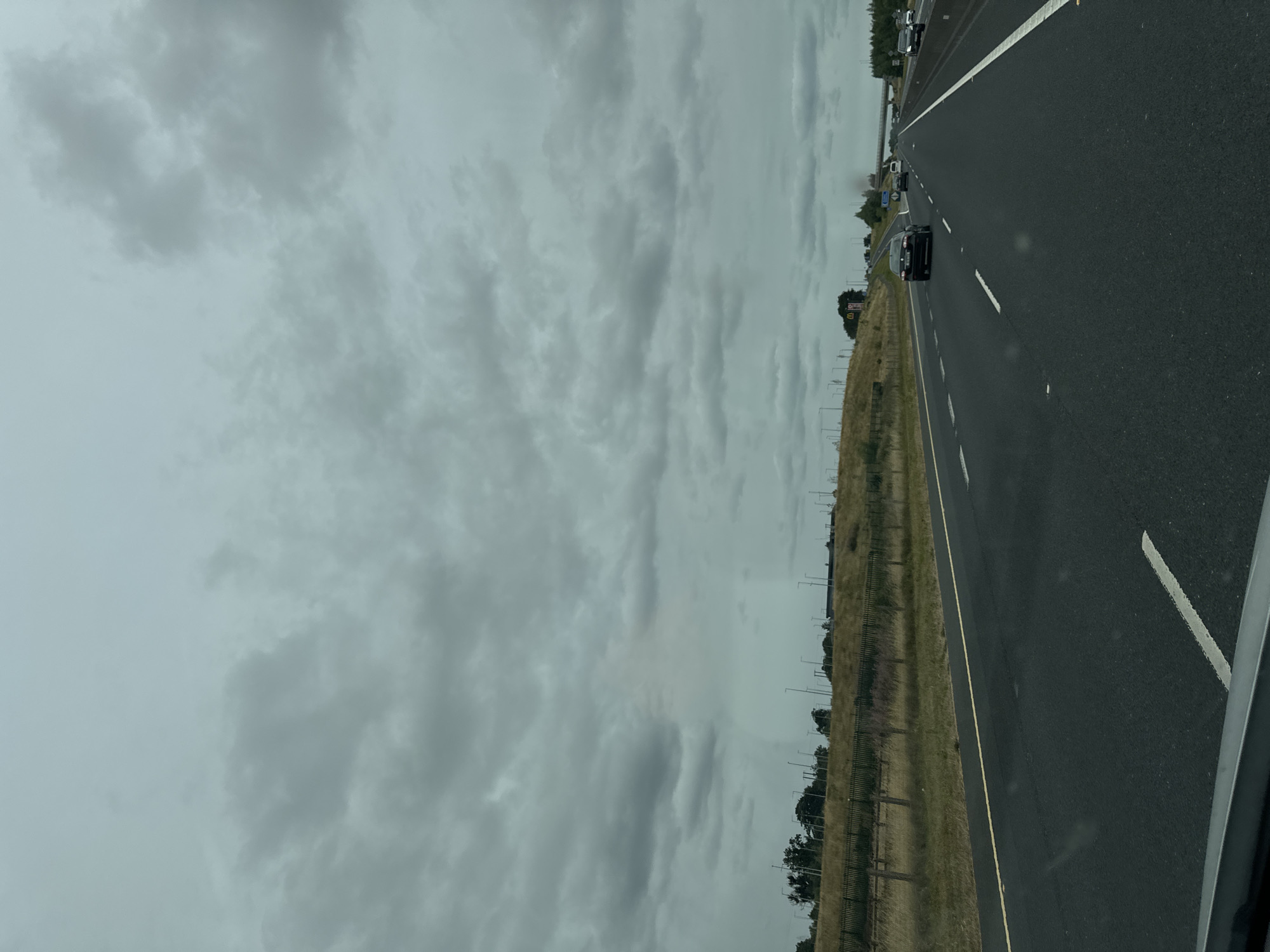The image shows a view of a highway in the vicinity of Kilgowan Cottages, Ireland, under a cloudy sky. The highway has multiple lanes, with cars driving in the same direction. A grassy embankment runs alongside the highway, with trees and lampposts visible in the distance. The sky is filled with dark, heavy clouds, suggesting an overcast or rainy day. The overall impression is one of a typical suburban or rural highway landscape.