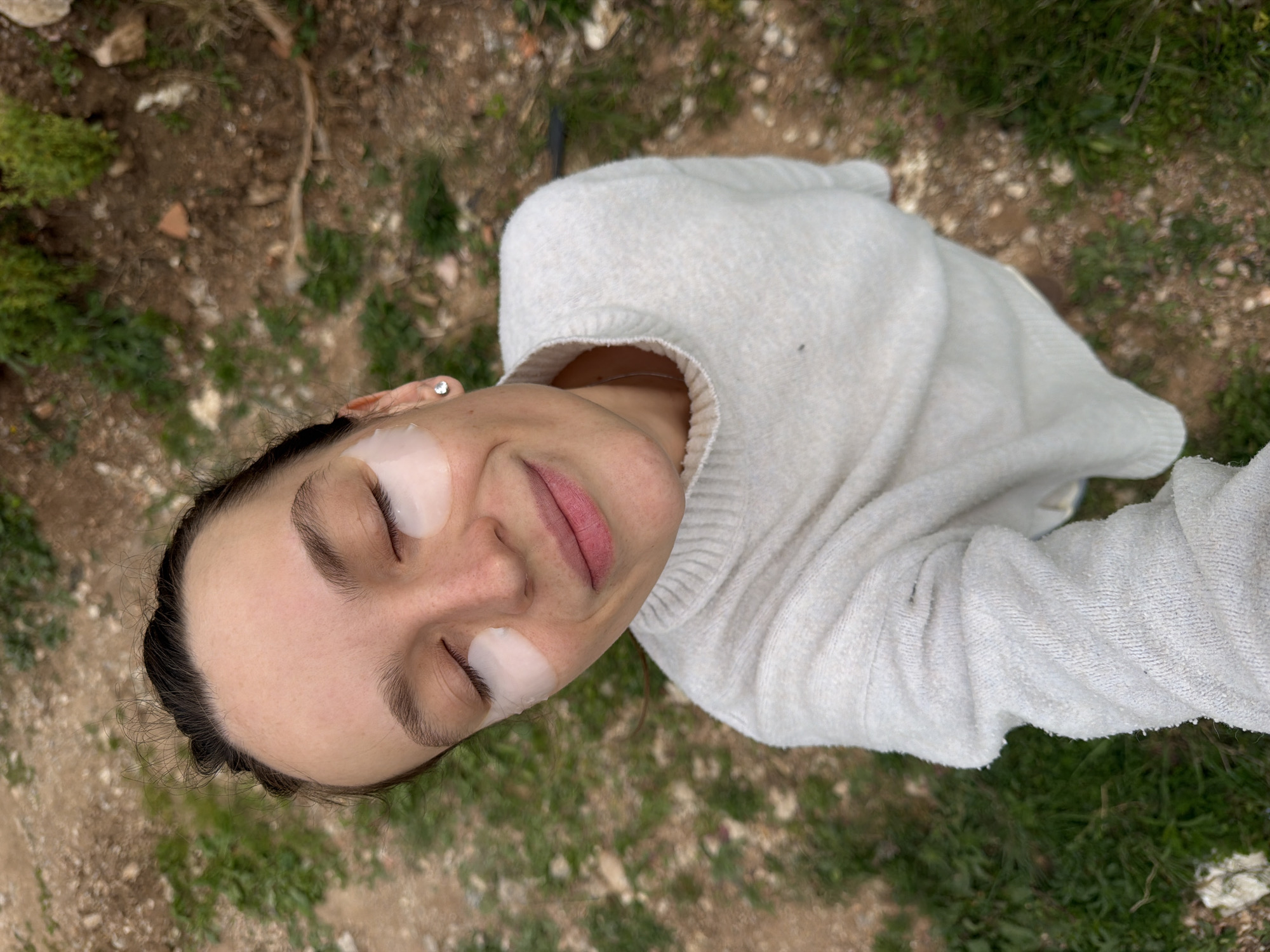A person is lying on a surface with grass and dirt. The person is wearing a white sweater and has eye patches under both eyes. The person's eyes are closed, and they are smiling. The background includes grass, dirt, and small rocks. Based on the general aesthetic and the person's attire, this image does not give enough details to be precise about the city of Budva, Montenegro.