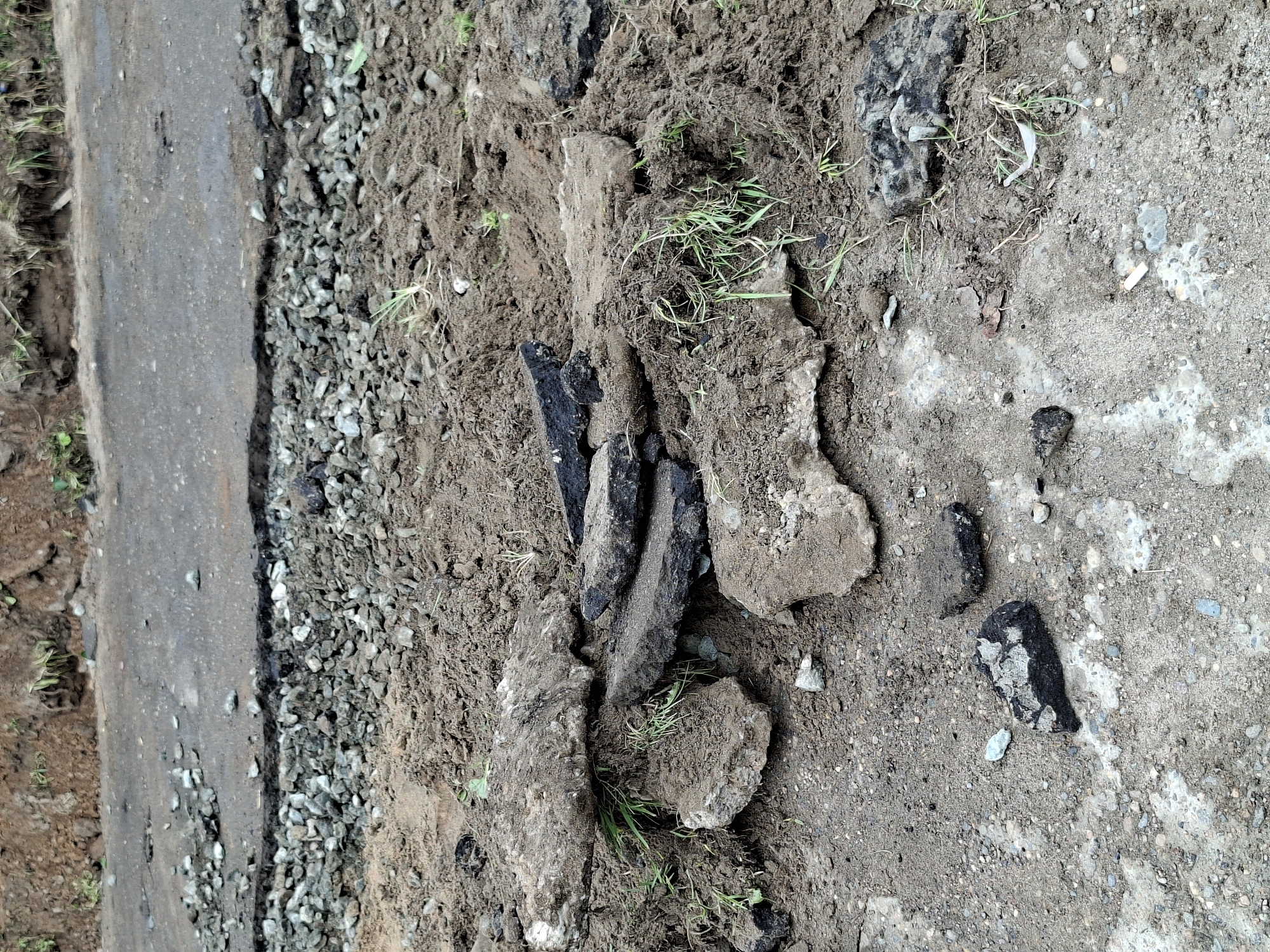 The image displays a close-up, top-down view of a section of ground featuring damaged or disturbed surfaces. On the left, a dark, narrow strip of weathered pavement, likely asphalt, runs vertically along the edge. Adjacent to this pavement is a band of light-grey gravel composed of small, irregular stones.
To the right of the gravel, the majority of the frame is covered by disturbed, dark brown soil and mud. This area is uneven, with numerous chunks of debris embedded within it. Some of these debris pieces are dark and angular, resembling broken asphalt, while others are lighter in color and more textured, appearing to be fragments of concrete or rock. Scattered patches of green grass and weeds are visible growing amongst the soil and debris, indicating some vegetation regrowth.
Further to the right, the ground becomes lighter in color and appears drier, with scattered small pebbles and an irregular pattern of a white, possibly powdery, residue on the surface. A small white cylindrical object, resembling a cigarette butt, can be seen on this lighter ground.
The scene suggests an outdoor environment, possibly a roadside, pathway, or construction area, where a paved surface has been broken up, eroded, or excavated, exposing various layers of ground material. The lighting indicates it is daytime, with no harsh shadows. No specific activity is taking place, but the state of the ground implies recent disturbance or ongoing environmental wear. There is no visible text in the image.