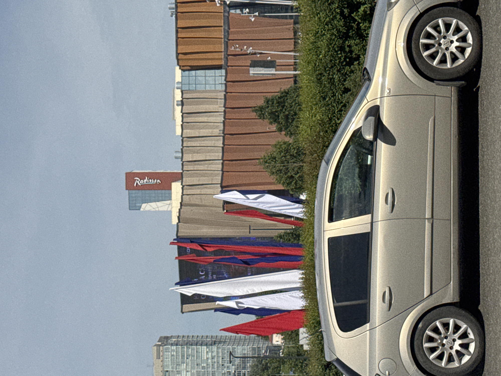 A sideways shot taken at eye-level shows a light grey car parked on a street in Perm, Russia. In the background is a collection of buildings including one displaying a "Radisson" sign. Red, white, and blue flags are seen attached to flagpoles in the foreground. Green shrubbery is visible near the car.