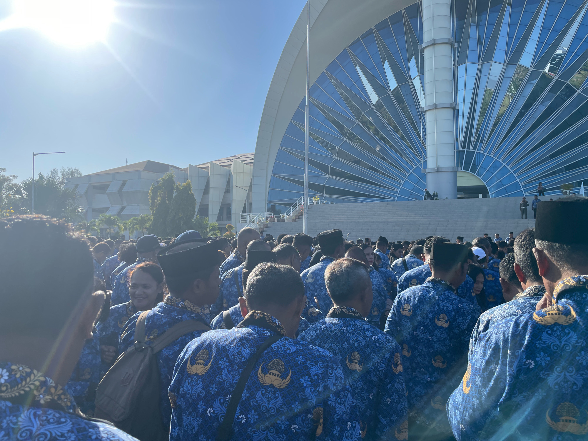 A large group of people, predominantly men wearing matching blue patterned uniforms with gold emblems, are gathered outdoors on a sunny day. The uniforms appear to be official attire. The building in the background is modern and features a distinctive arched glass facade. Some individuals are looking towards the camera, while others are looking in different directions, suggesting a formal gathering or event. One woman in the crowd is smiling and looking towards the camera. The scene is set against a bright blue sky with the sun prominently visible and casting a bright glare. The foreground shows the backs of several individuals in the uniform, emphasizing the collective nature of the gathering. The overall impression is one of an organized event involving government or public sector employees. The building's unique architecture suggests it could be a significant landmark. It is highly likely that this scene is located at the Gubernur NTT Building, located in Kupang, Indonesia, as it features the architectural style and emblem consistent with governmental institutions in the region.