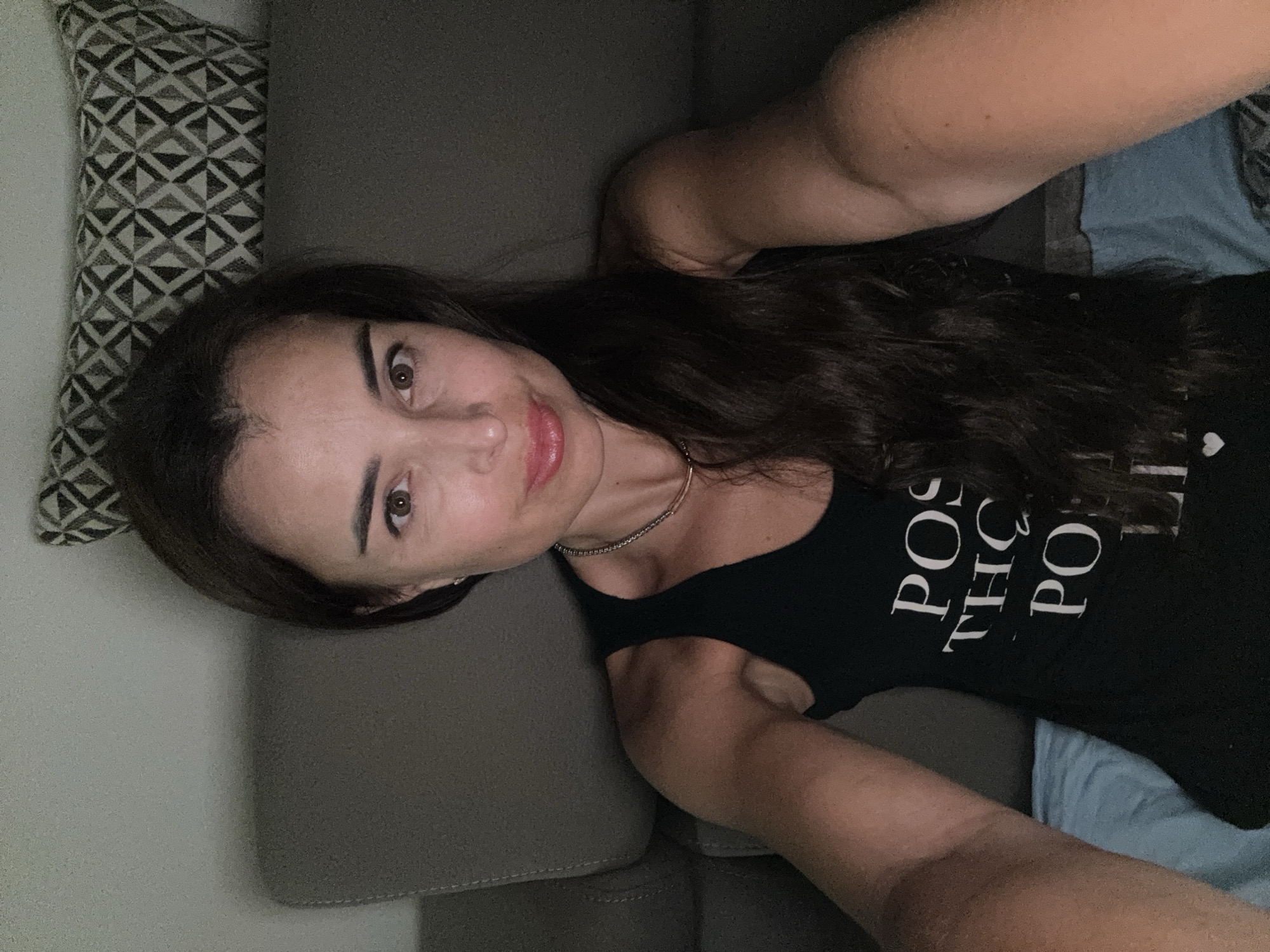 A woman is seen from the chest up, taking a selfie. She has long, dark hair that falls over her right shoulder and down her back. She is wearing a black tank top with white text that reads "POS THE PO... LI...". She has light skin and brown eyes, and she is wearing a thin silver necklace. The background appears to be a couch with a patterned pillow. The lighting is dim, suggesting the photo was taken indoors at night.