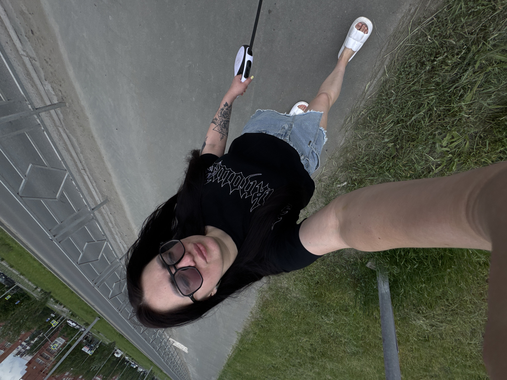 A top-down, high-angle shot shows a woman standing on a sidewalk next to a grassy area. She wears a black shirt, a denim skirt, and white sandals. She is holding a retractable dog leash in her left hand, and her right arm extends towards the camera, suggesting she is taking a selfie. A tattoo is visible on her left arm. She also wears eyeglasses. A gray road is visible behind a low metal fence to the left, with buildings and cars in the distance. The location is outdoors and appears to be in a city.
