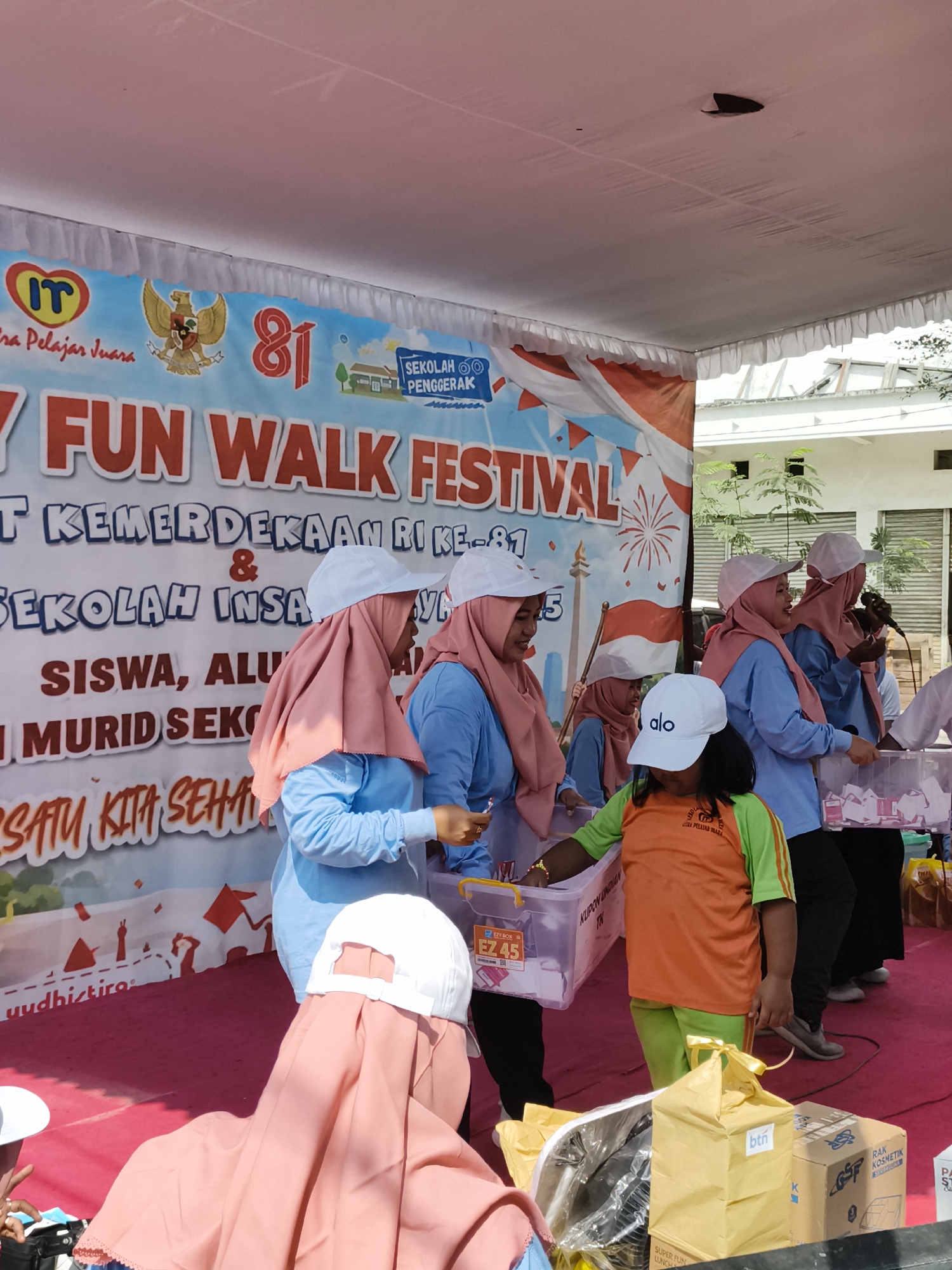 The image depicts an outdoor event in Petiken, Indonesia, likely a school-related activity given the banners and the presence of uniformed individuals. In the background, a large banner displays the text "Y FUN WALK FESTIVAL" and mentions "SEKOLAH INSA" and "SISWA, ALUMNI, MURID SEKOLAH" suggesting a school festival commemorating Indonesian Independence Day ("KEMERDEKAAN RI KE-81"). The banner also features national symbols like the Garuda Pancasila emblem and the Indonesian flag.
In the foreground, several individuals are visible. Four women wearing pink hijabs and light blue shirts are standing behind a table or stage area. One woman is speaking into a microphone, while others are interacting with clear plastic bins containing what appear to be raffle tickets or small items. Another young person in an orange and green shirt and a white cap is also interacting with a bin. Several white caps are visible on individuals' heads, some with "alo" printed on them.
On the table or stage, there are several boxes, including a yellow gift-wrapped box with "btn" visible, and a cardboard box labeled "GSF RAK KOSMETIK." The overall atmosphere suggests a community event, possibly a fundraising or celebratory gathering, taking place under a white canopy with ruffled edges. The ground is covered with a red carpet.