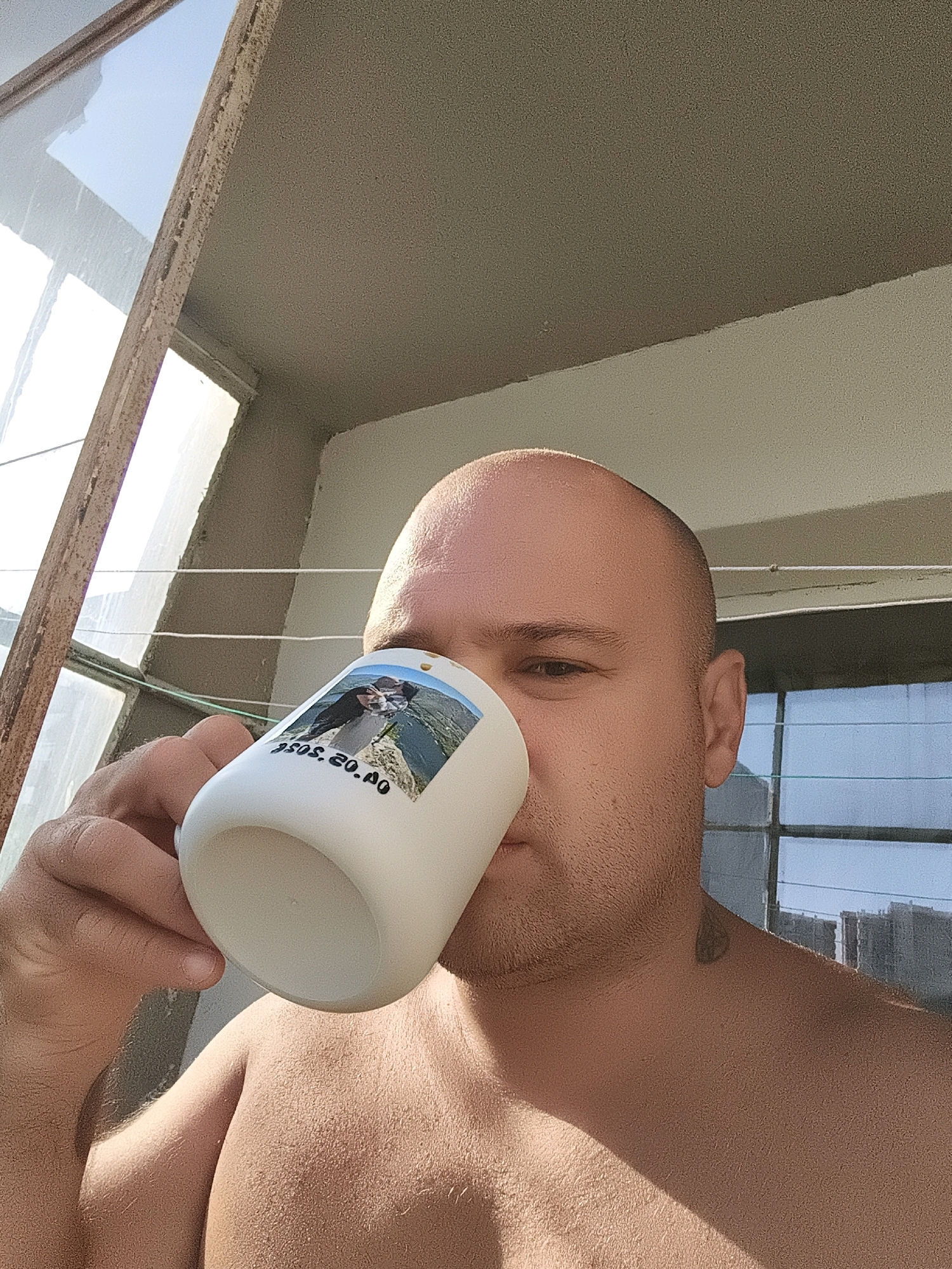 An outdoor, low-angle, medium shot shows a bald man holding a white mug to his lips. The mug has a picture of a couple and the date "1958.20.10" printed on it. The man is shirtless, and his bare shoulder and chest are visible. He has a light beard and a tattoo on his neck. The background features a window frame and what appears to be a balcony with some washing lines. The lighting suggests it is daytime, possibly morning or afternoon, with strong sunlight casting shadows. The overall environment appears to be urban, but the specific location is not identifiable.