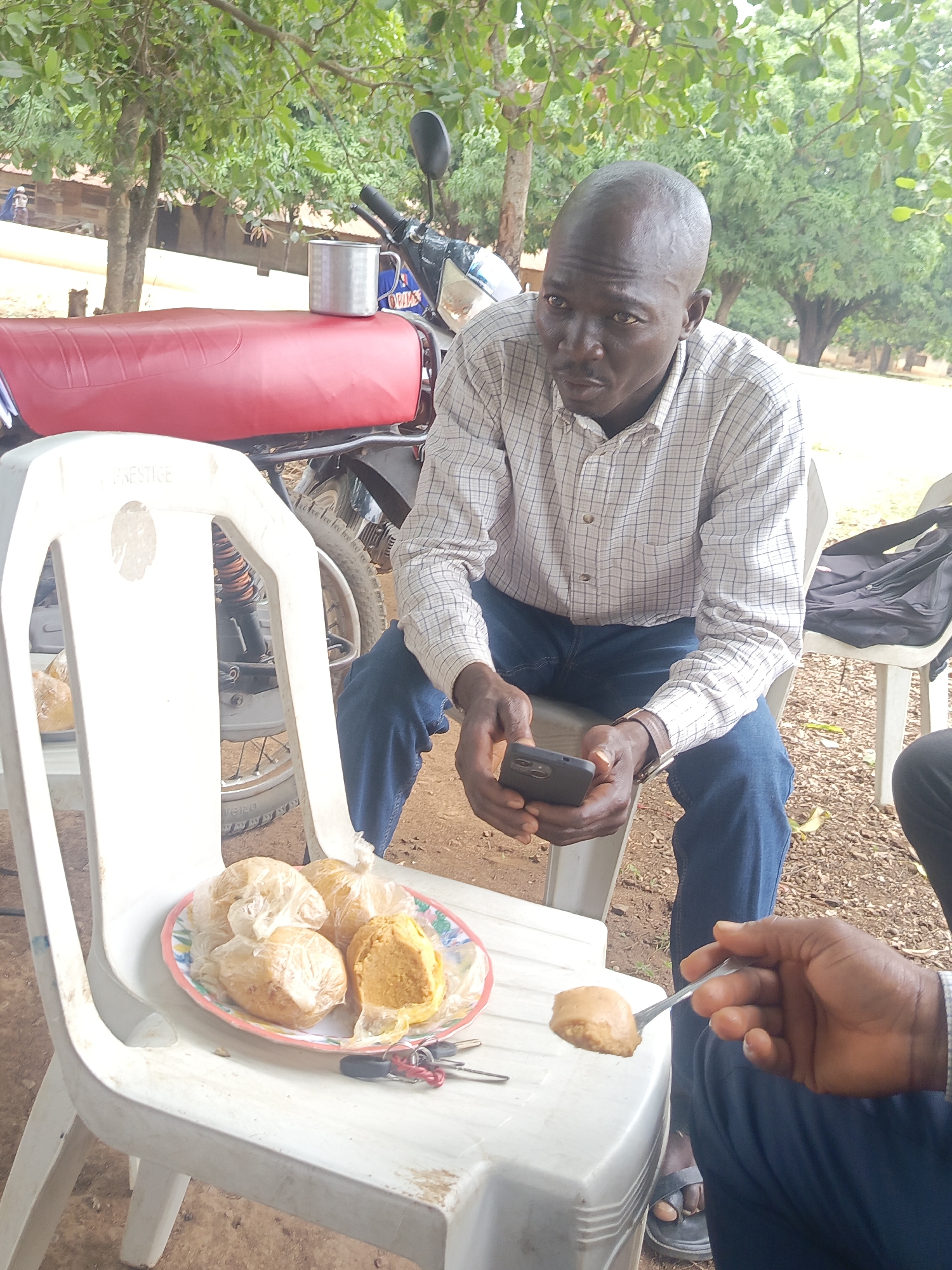 The image captures an outdoor scene in Andaha, Nigeria, depicting two individuals engaged in a casual activity under the shade of lush green trees.
In the foreground, a dark-skinned man with a bald head, wearing a light-colored plaid long-sleeve shirt and blue jeans, is seated on a white plastic chair. He holds a black smartphone in both hands and looks with a neutral expression slightly to his left. To his right, only the lower arm and hand of another person are visible, holding a metal spoon with a portion of brownish-yellow food. This person is also wearing dark trousers and black sandals.
Between the two individuals, a third white plastic chair holds a plate of food. The plate contains several portions of a yellowish-orange, textured food, three of which are wrapped in clear plastic, while one is unwrapped. A set of keys with red and black fobs rests beside the plate. The brand name "PRESTIGE" is visible on the backrest of this chair.
In the mid-ground, behind the prominent man and the chairs, a motorcycle with a distinctive red seat is parked. A silver metal mug sits on the motorcycle's seat. In the background, simple structures or buildings can be seen through the dense foliage of the trees. The ground is a mix of dirt and gravel. The bright, yet diffused lighting suggests it is daytime, possibly late morning or early afternoon, with comfortable weather under the tree cover.