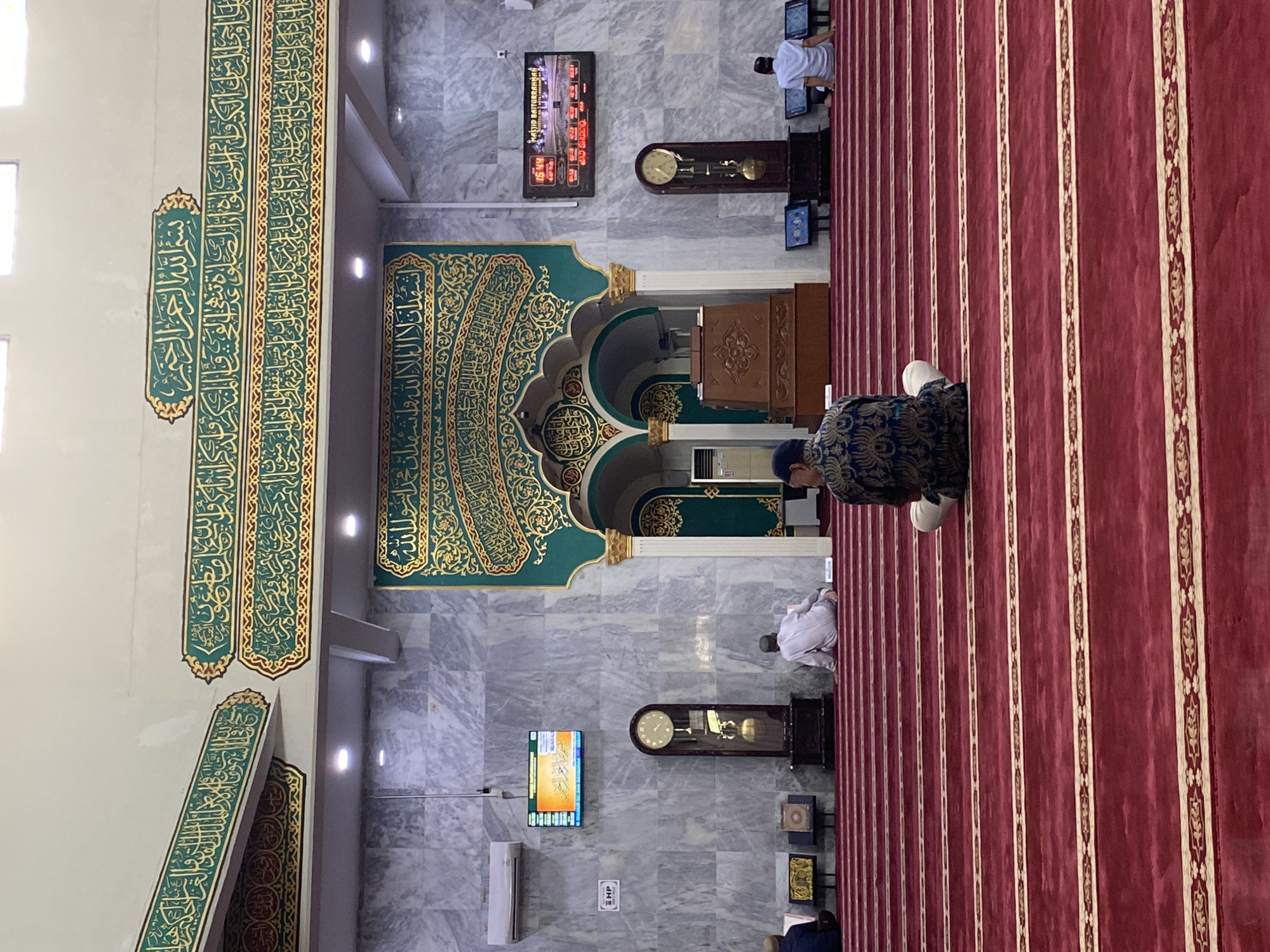 The image depicts the interior of a mosque in Lhokseumawe, Indonesia, identified by the text on the digital display as "MASJID BAITURRAHMAN LHOKSEUMAWE." The scene is viewed from a low angle, facing the front of the prayer hall. A person wearing a blue and gold patterned tunic and white pants is seated on the red carpeted floor, facing away from the camera and towards the mihrab area. The carpet is deeply red with alternating stripes of gold ornamentation.
In the background, the mosque's architecture is adorned with intricate green and gold Arabic calligraphy displayed prominently on the walls. To the left, a large grandfather clock with a brass pendulum stands against a marble-patterned wall. A digital display screen is mounted above the clock, showing prayer times and the mosque's name. To the right, another digital display shows similar information. In the distance, several other individuals are seen seated on the prayer mats, some engaged in prayer or contemplation. The mihrab, the semicircular niche indicating the direction of prayer, is visible in the central background, framed by ornate arches. The environment appears clean and well-maintained, suggesting it is an active place of worship.