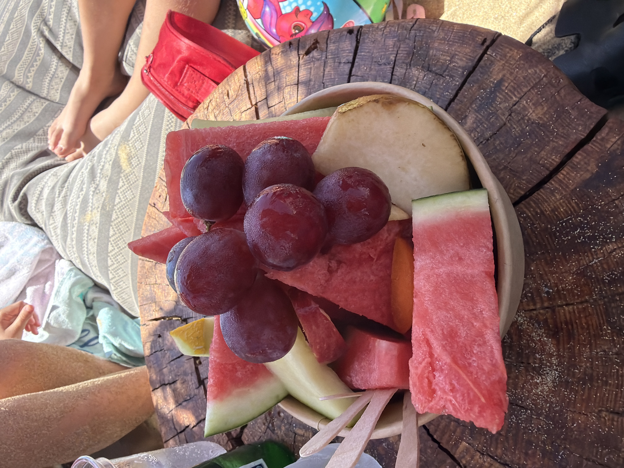 The image depicts a bowl of fruit on a wooden surface. The fruit includes watermelon, grapes, pear or apple, and melon. Wooden forks stick out from the bottom of the bowl. The wooden surface appears to be a section of a tree trunk. Sand covers the surface. To the left, a person sits with a striped blanket. A red purse rests on the blanket. In the background are legs of other people and a beach ball. Based on the provided information, the location is Nea Peramos, Greece.