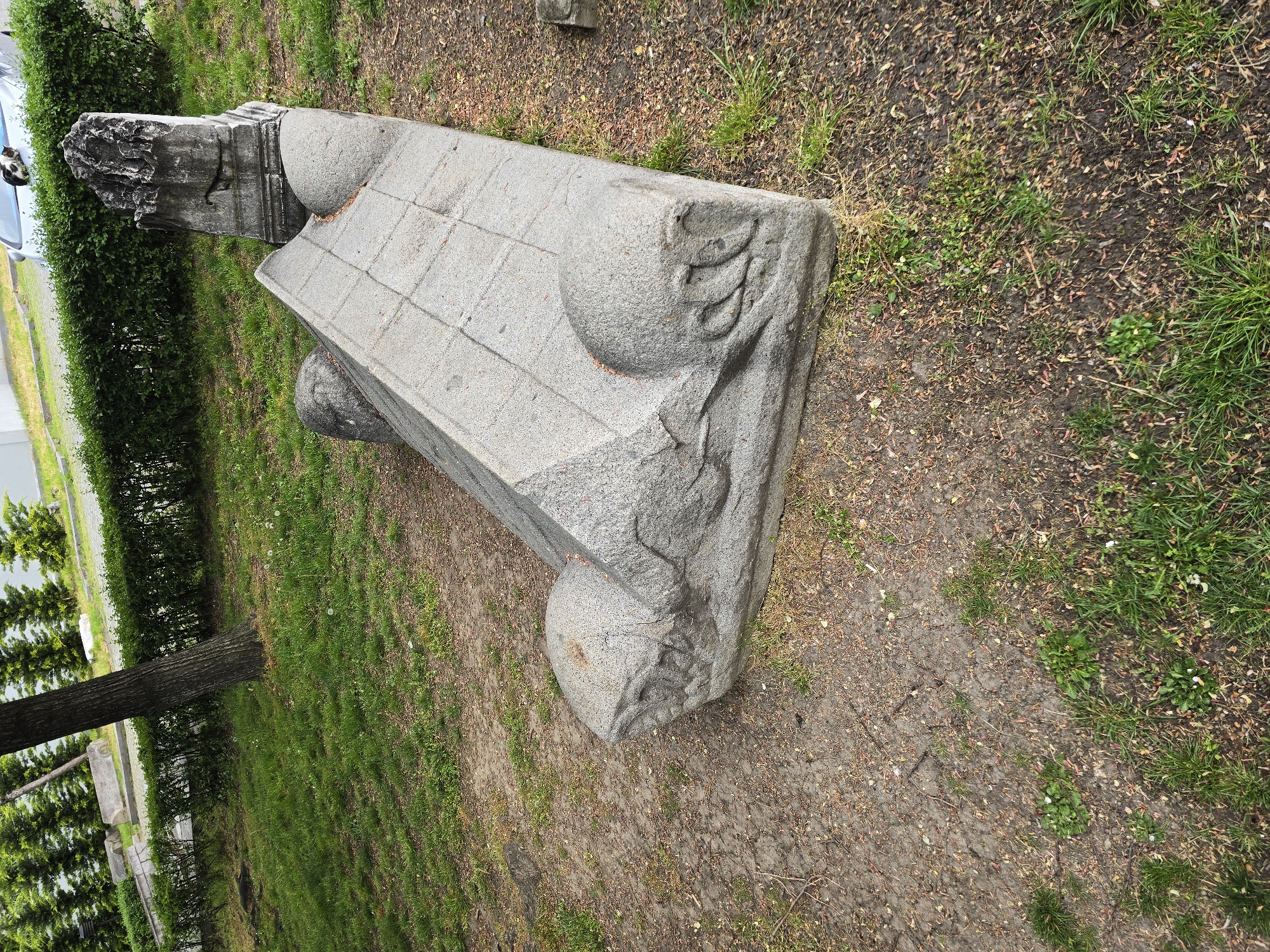 The image shows a large, ornate stone structure, likely a fragment of a building or monument, resting on the ground in a grassy area. The structure is composed of large, rectangular blocks of gray stone with rounded, bulbous protrusions and carved details. The stone surface is visibly aged and weathered. The surrounding environment consists of grass, dirt, and sparse vegetation. In the background, there is a row of green bushes and a glimpse of a building or vehicle. The location is in the city of Unknown, Unknown.