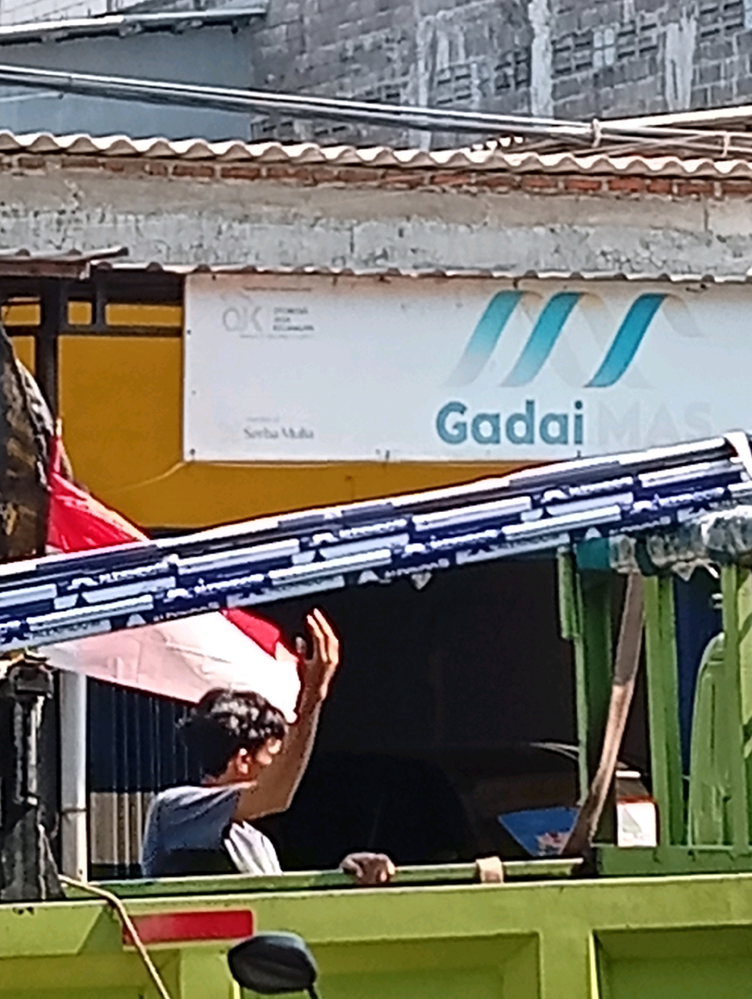 In Bangah, Indonesia, a young man is seen in the back of a green truck, holding a white and red Indonesian flag. He is wearing a blue t-shirt. The truck has a long, blue-and-white striped railing on top. In the background, a building with a yellow wall and a white sign with blue lettering that says "Gadai MAS" is visible. Part of a brick wall is also visible above the building. The scene appears to be outdoors on a sunny day.