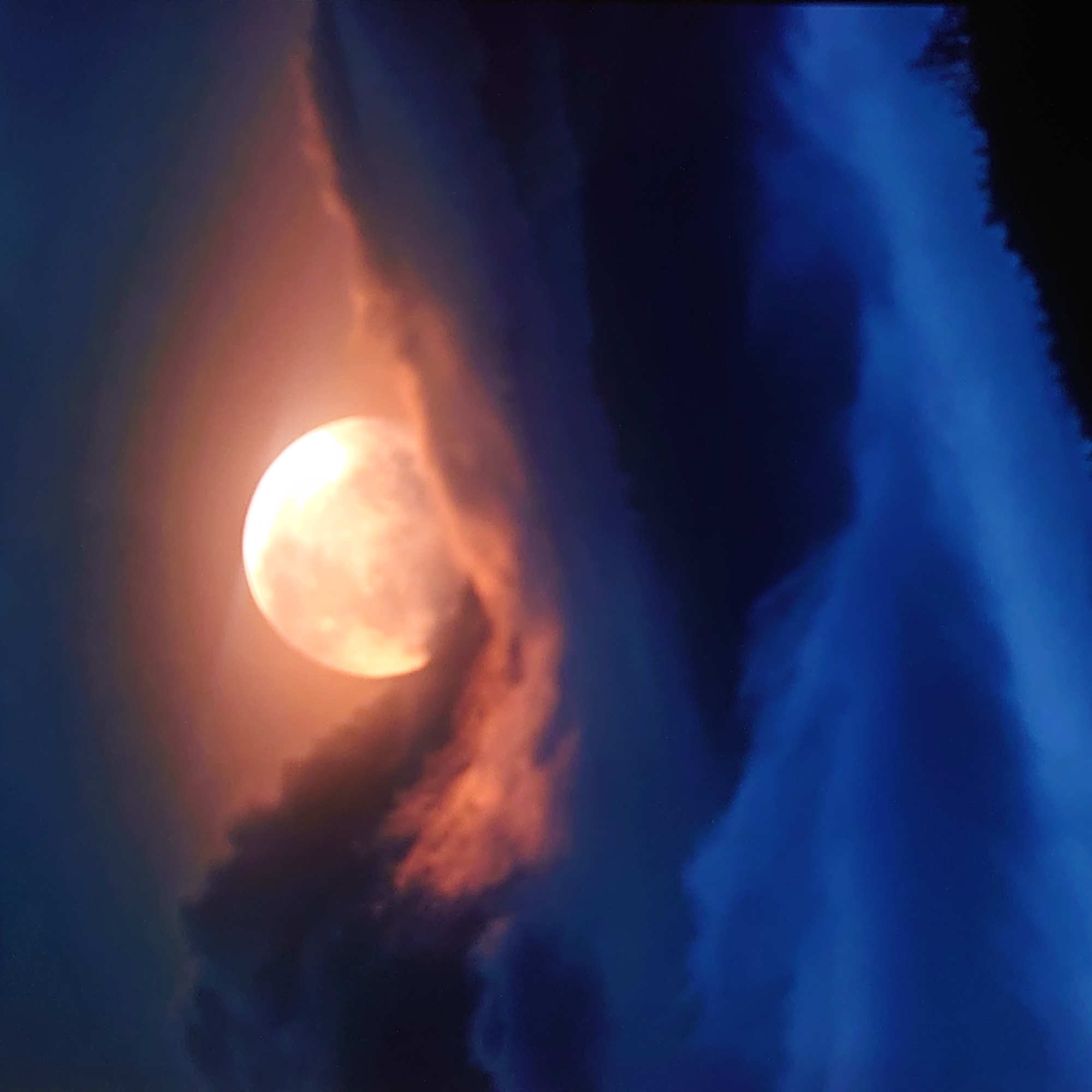 The image depicts a full moon glowing with an orange hue, partially obscured by dark, textured clouds tinged with pink and orange light. Below the moon, layers of deep blue clouds form a misty expanse, creating a sense of depth. The lower part of the image shows a dark silhouette of a treeline against the blue of the clouds and the sky. There are no people or specific urban elements visible to indicate a location in Belorechensk, Russia. The scene is a natural landscape under moonlight and cloud cover.