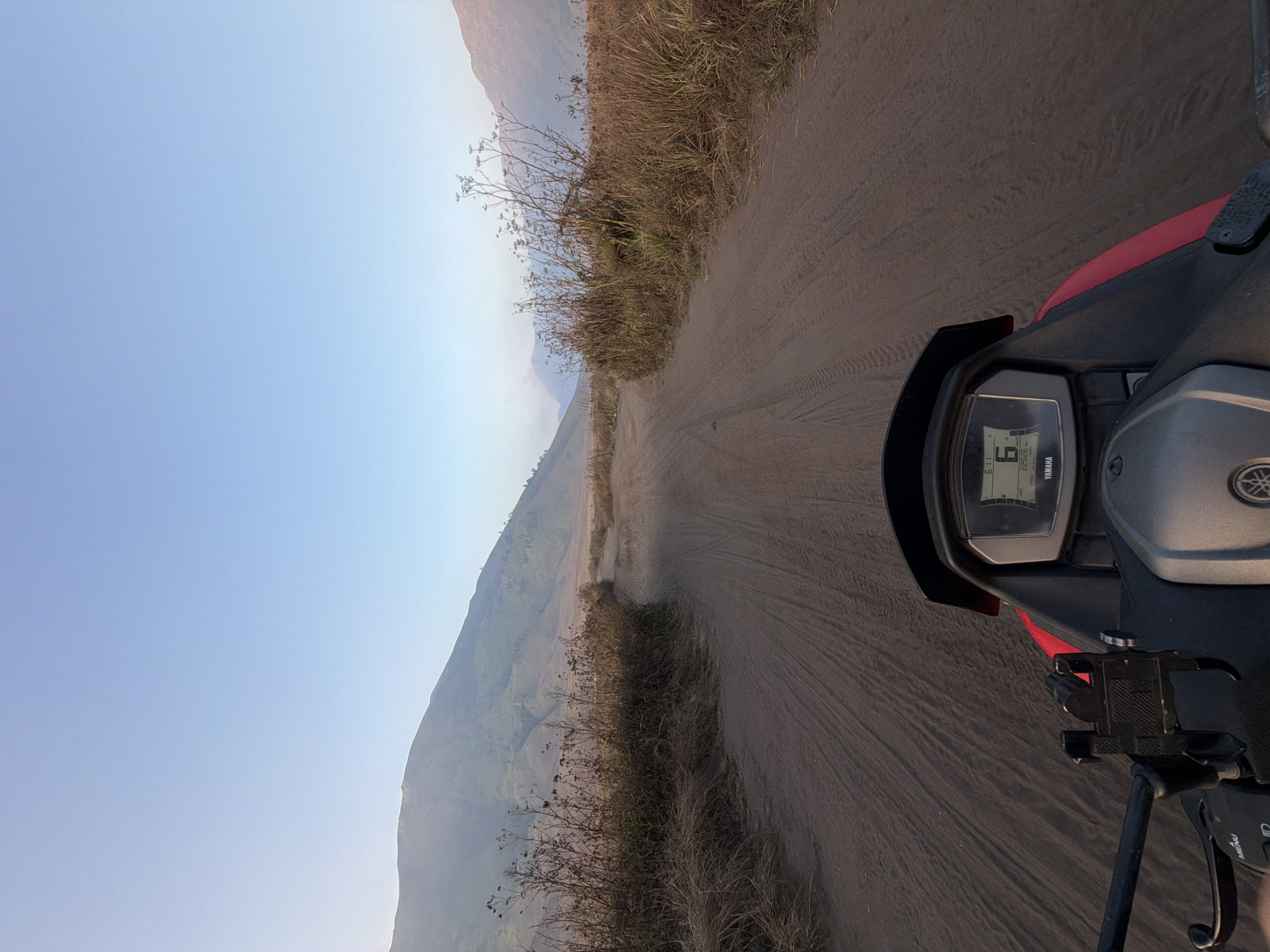 A first-person point-of-view photograph captures a dusty, dirt road stretching through a barren landscape. The road is marked with tire tracks, suggesting recent travel by a vehicle. Dry, brown vegetation lines both sides of the road, with a large, undulating mountain range in the background. The sky above is clear and a soft blue, indicating either early morning or late afternoon. In the foreground, the dashboard of a red motorcycle is visible, displaying a digital speedometer reading "6" and "22926". The motorcycle also has a phone mount attached to the handlebars. The scene appears to be located in a rural or natural environment, possibly a savanna or desert region, given the sparse vegetation and the presence of mountains. The overall impression is one of travel and exploration in a remote, natural setting.