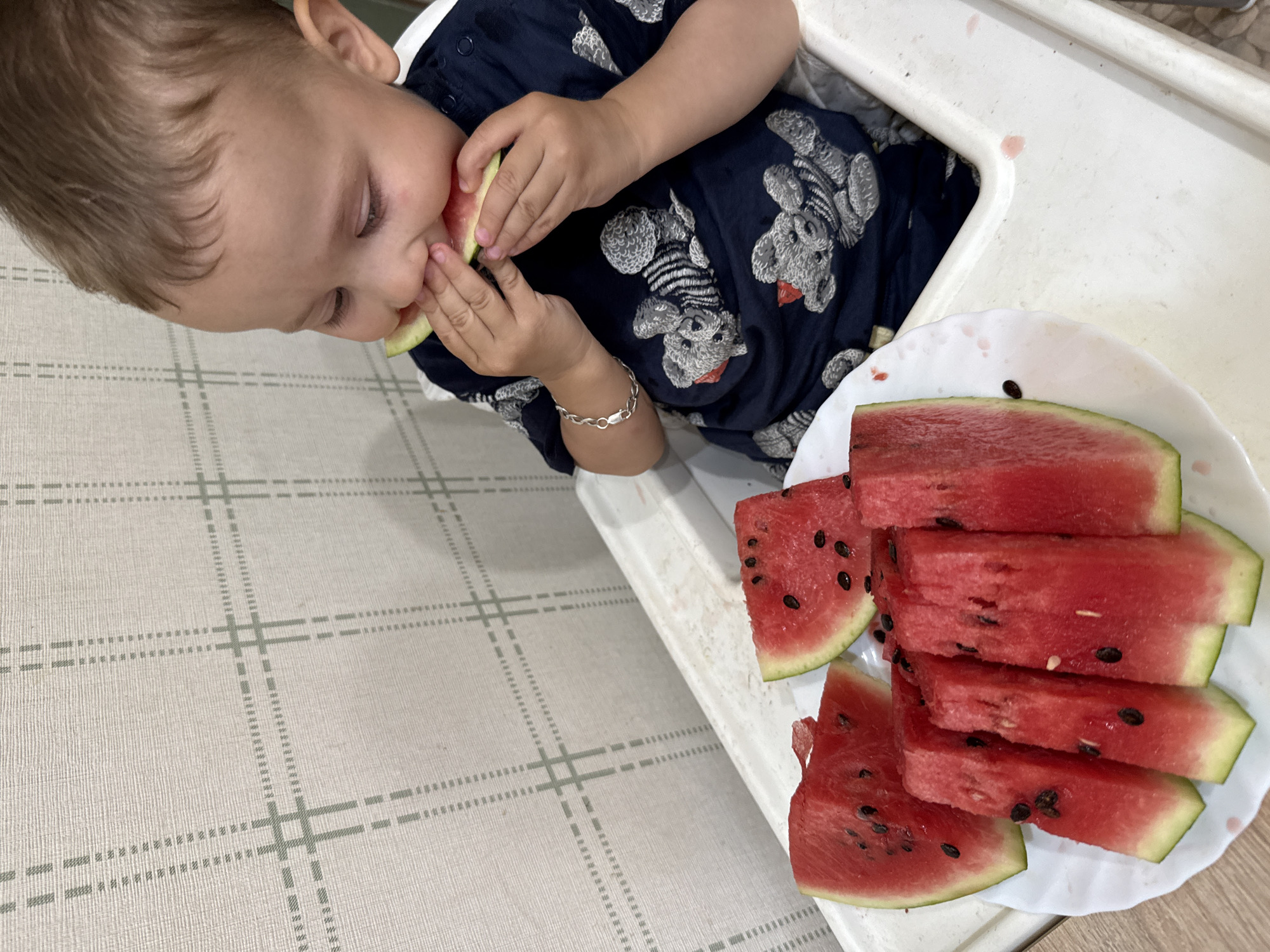 An overhead-angle indoor photograph depicts a young male toddler seated in a white plastic high chair, actively eating a slice of watermelon. The child, with light brown hair and fair skin, wears a dark blue short-sleeved shirt patterned with stylized grey and white teddy bears, and a silver-colored chain bracelet on his left wrist. He holds a triangular piece of watermelon to his mouth with both hands, concentrating on biting into the red flesh. To the child's immediate left, on the high chair's white tray, a white plate with a scalloped edge holds multiple additional slices of red watermelon, some triangular and some elongated rectangular, all containing black seeds and showing green-white rinds. The high chair tray exhibits several small pinkish-red stains, consistent with watermelon juice. The background consists of a light beige or off-white wall covered in wallpaper featuring a repeating grid pattern composed of light grey-green solid, dashed, and dotted lines, indicating an indoor residential setting in the city of Unknown, Unknown.