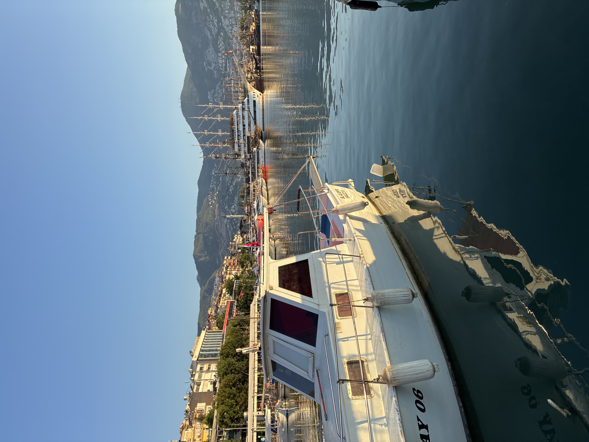 In Alanya, Turkey, a peaceful morning scene unfolds in the harbor. The sun, low on the horizon, casts a warm glow across the water and the docked vessels. In the foreground, a white motorboat with red-tinted windows is tethered, its reflection shimmering on the calm, blue surface of the sea. The boat is marked with the number "06". Behind it, a collection of larger, multi-masted sailing ships, some appearing historic or replica, are moored. The shoreline is lined with buildings of varying architectural styles, from modern structures to older, colorful houses, all nestled against a backdrop of lush green hills that rise to form the distant mountains. The sky is a clear, bright blue, indicating a pleasant day ahead. There are no people visible in the image. The overall atmosphere is serene and tranquil, capturing a quiet moment in the bustling port.
