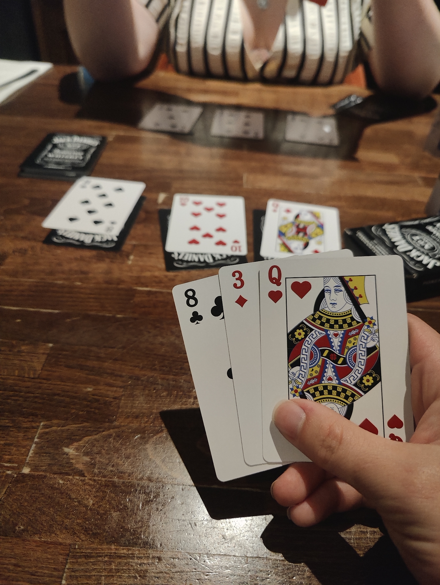 A close-up indoor shot shows a person holding three playing cards: 8 of Clubs, 3 of Diamonds, and Queen of Hearts. The cards are held in a hand, with the Queen of Hearts facing the viewer. Several other playing cards are visible on a wooden table in the background. Some of the cards on the table are also visible, as are several Jack Daniel's playing card decks. The location of the image is not specified, and there is no information indicating it is in East Suffolk, United Kingdom.