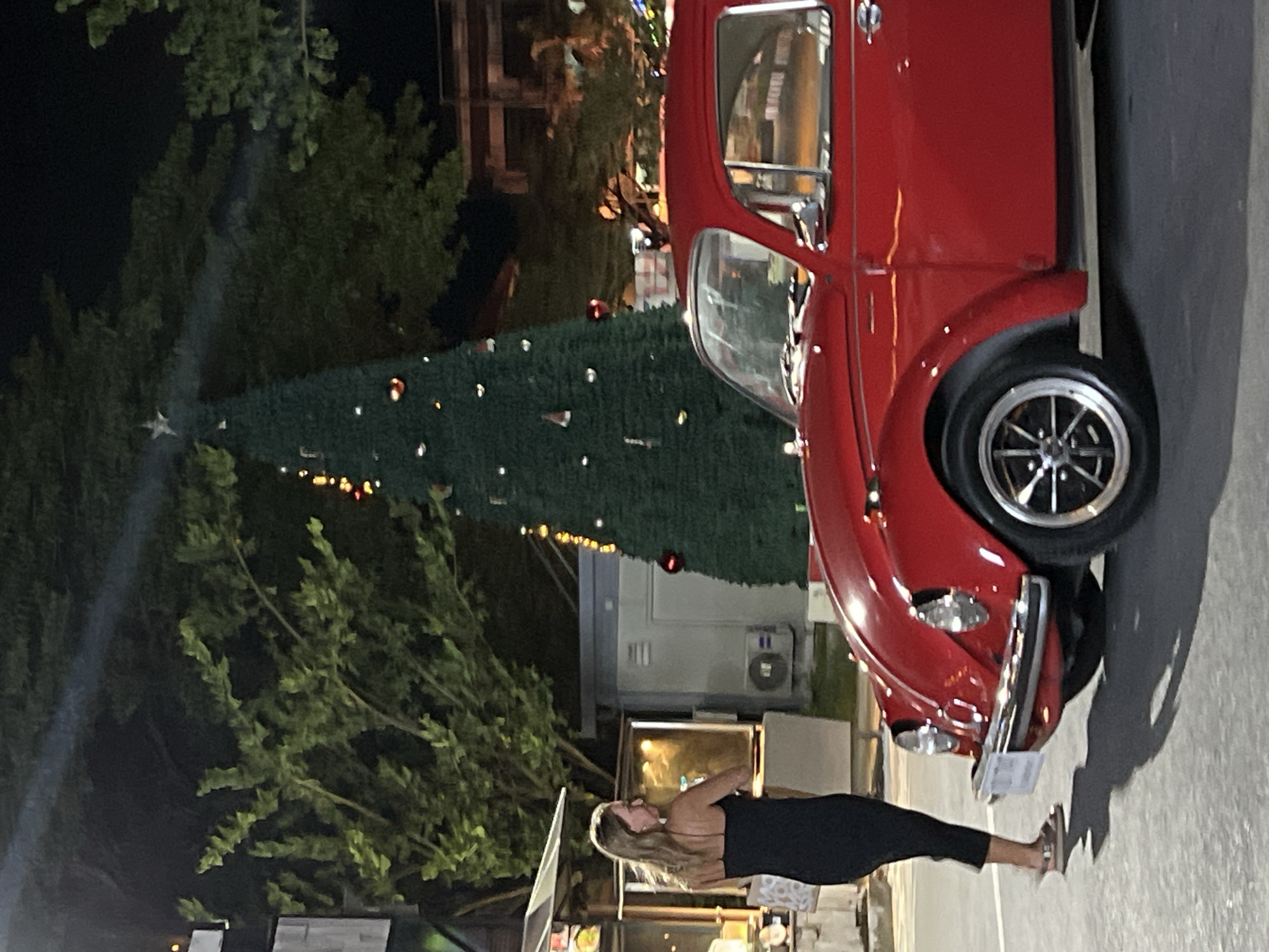 At night in Choeng Thale, Thailand, a woman in a black dress stands next to a red Volkswagen Beetle. Behind them, a decorated Christmas tree is illuminated. The woman is facing away from the camera, looking to her left. The car is parked, and its headlights are on. The background includes trees, buildings, and streetlights. The ground is paved.