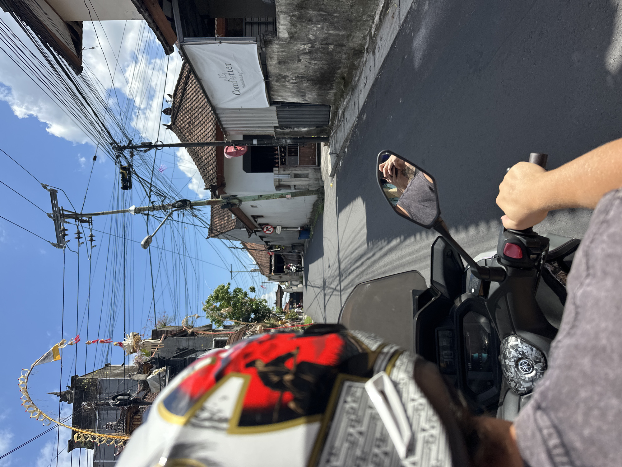 The image depicts a first-person perspective of someone riding a scooter down a narrow street in North Kuta, Indonesia. The rider is wearing a helmet with a red, white, and black design, and their arm and hand are visible on the handlebars. A mirror on the scooter reflects the rider's upper body and the road ahead. The street is paved and lined with buildings, some of which have traditional Balinese architecture with decorative roofs. Overhead, a dense network of electrical wires is strung across poles, common in many parts of Indonesia. Several decorative arches and banners, likely for a local festival or ceremony, are visible on the left side of the street, attached to buildings and poles. The sky is blue with scattered white clouds. Further down the street, other people on scooters and a few pedestrians can be seen. A white banner with text, possibly advertising, hangs on the right side of the street. The overall scene suggests a typical day in a bustling Indonesian town.