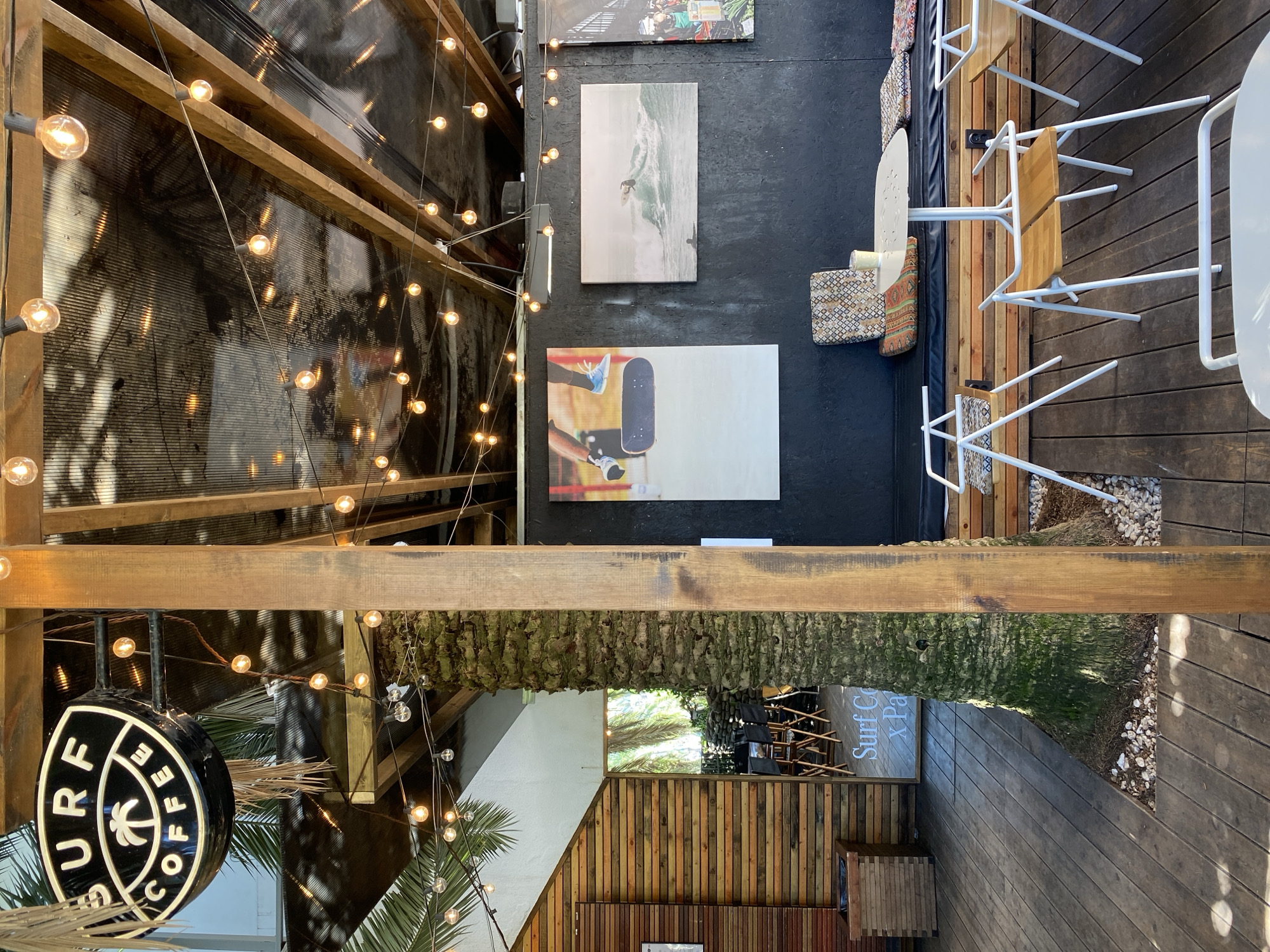 This image depicts an outdoor patio area of Surf Coffee X Park, a cafe in Sochi, Russia. The view is from an elevated perspective, looking down.
On the left side, there is a structure with wooden beams and string lights, casting a warm glow. A round sign for "Surf Coffee" is visible, hanging from the structure. Palm trees are also present, adding to the tropical atmosphere.
The center of the image features a black area with artwork displayed on the wall. One artwork depicts a surfer riding a wave, while another features a skateboard. There are also tables and chairs with white frames, arranged on a wooden deck. Decorative cushions are placed on some of the seats.
On the right side of the image, there is a continuation of the wooden deck, along with a view of a palm tree and a sign for "Surf Coffee X Park". The overall environment appears to be a relaxed and inviting outdoor space, with a surf-themed aesthetic.