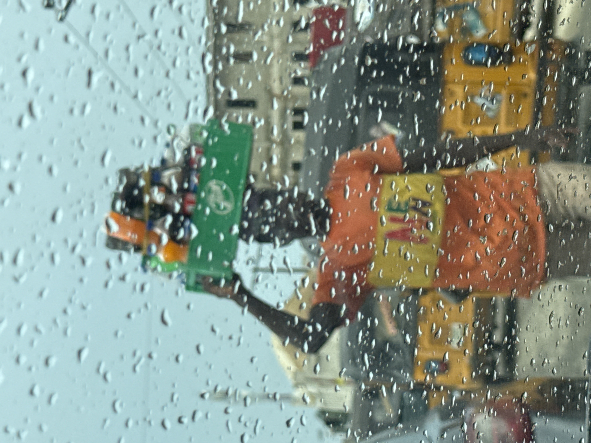 A street vendor in Ado, Nigeria, is seen through a rain-streaked window. He holds a green plastic basket containing bottles. He wears an orange shirt with yellow detail. Yellow containers are stacked behind him. The background includes blurry buildings.