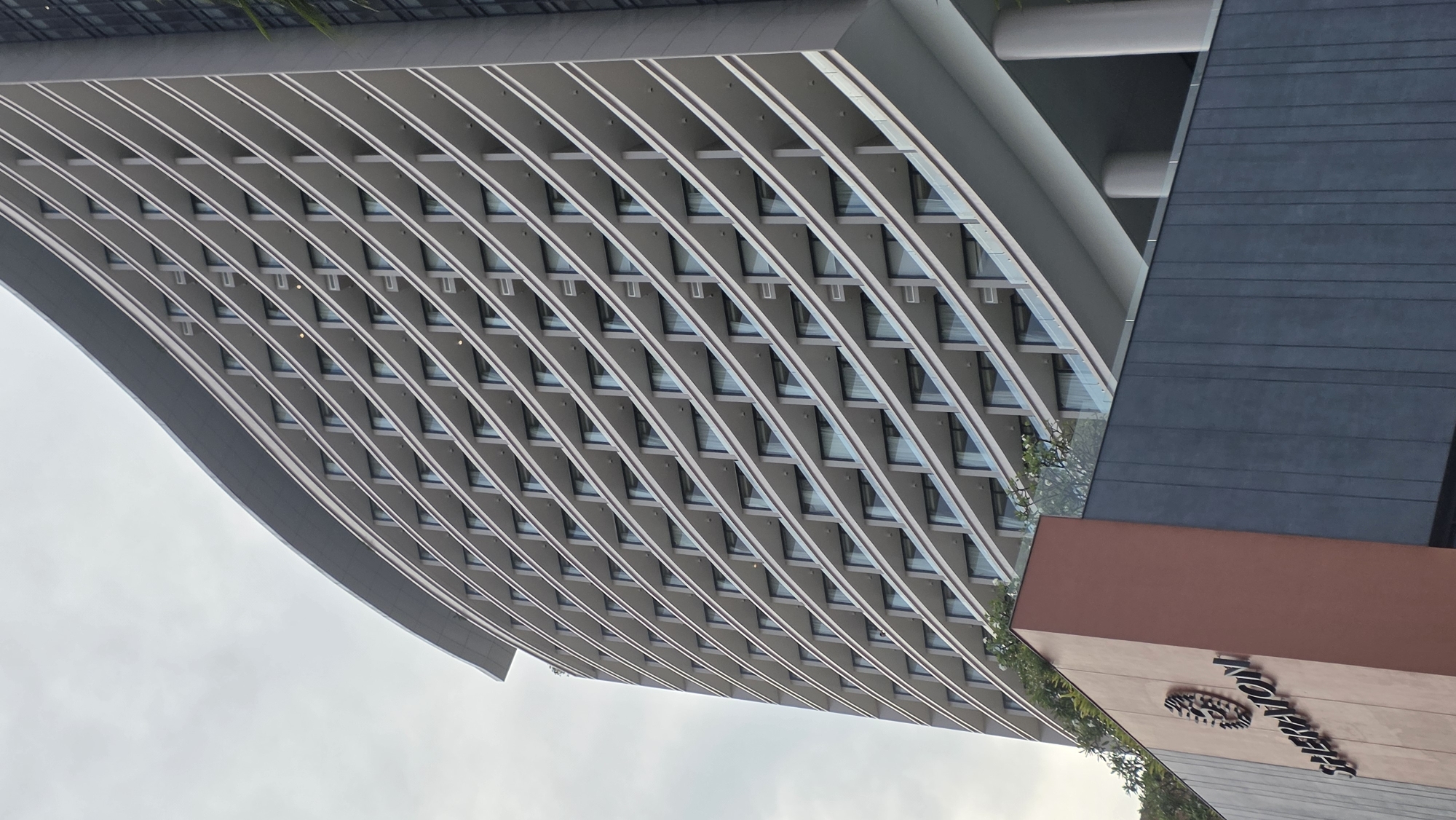 An outdoor, upward-angled shot captures the modern architecture of a large building complex, likely a hotel or resort, under an overcast sky. The primary structure on the left features a distinctive, multi-story facade characterized by numerous curved horizontal lines, forming a wave-like pattern of balconies or sunshades. Each level contains multiple recessed windows, suggesting individual rooms or units within. To the right, an adjacent building section is visible, presenting a contrasting dark grey, textured facade with horizontal grooves on its upper portion, and a solid reddish-brown lower section. On this reddish-brown section, the word "SHERATON" is prominently displayed in a stylized white font, along with a partial view of its distinctive "S" logo, clearly identifying the building. Sparse green foliage and white flowering plants are visible near the base of the main building and around the signage area. The architectural style is contemporary, emphasizing clean lines and a unique, curvilinear design, indicative of a modern resort in a location like Nha Trang Ward, Vietnam. No specific activity or people are visible.