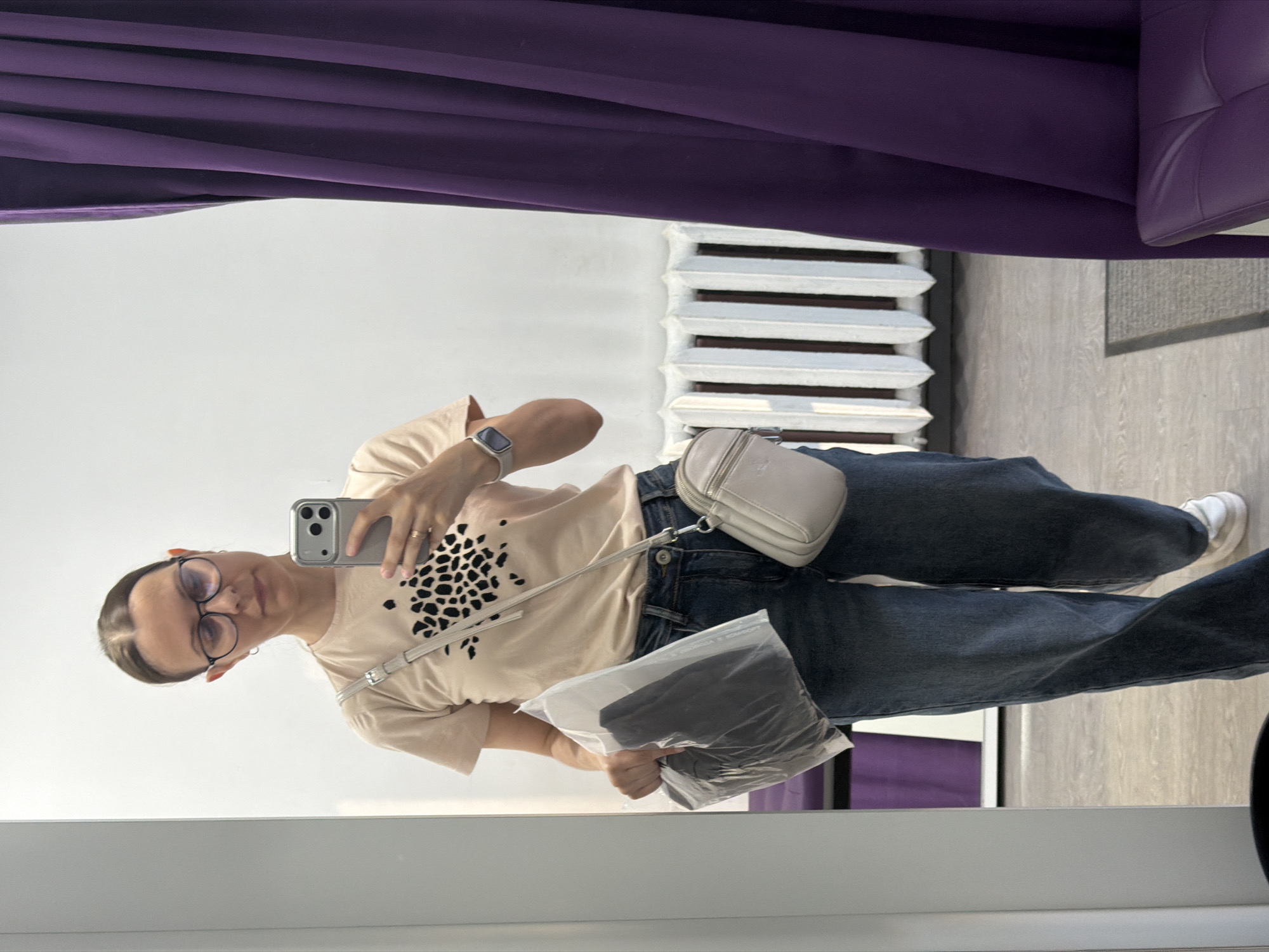 A woman is taking a mirror selfie in what appears to be a fitting room. She is wearing a light beige t-shirt with a black leopard print design, blue denim jeans, and white sneakers. She has a small, light beige crossbody bag and is holding a clear plastic bag containing dark clothing. She is also wearing glasses and a smartwatch. The background shows a white wall, a radiator, and a purple curtain. The floor is light gray laminate. The image is taken in Izhevsk, Russia.