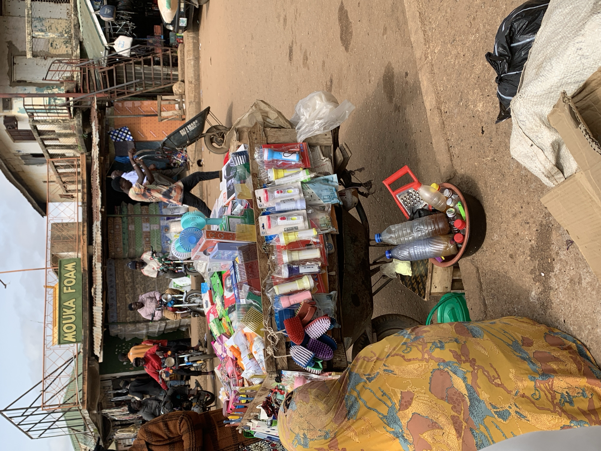 The image depicts a bustling street market scene in Bukuru, Nigeria. In the foreground, a woman wearing a yellow and brown patterned headscarf and a yellow patterned dress is partially visible from the back, facing away from the camera. To her right, a cart laden with various goods, including plastic combs, small fans, and flashlights in packaging, is prominently displayed. Some of the flashlights are red, white, and blue. Behind the cart, a man in a tie-dye shirt and black pants is pushing a wheelbarrow.
In the background, several people are seen on motorcycles. Three men are seated on motorcycles, one in a red top, another in a pink top, and a third in a white and red top. A sign overhead reads "MOUKA FOAM" in green letters against a yellow background. The architecture suggests a mix of commercial buildings with upper floors and awnings. The street is paved with dirt and gravel, and there are piles of garbage bags and cardboard boxes on the ground. The overall impression is one of a vibrant, active marketplace.