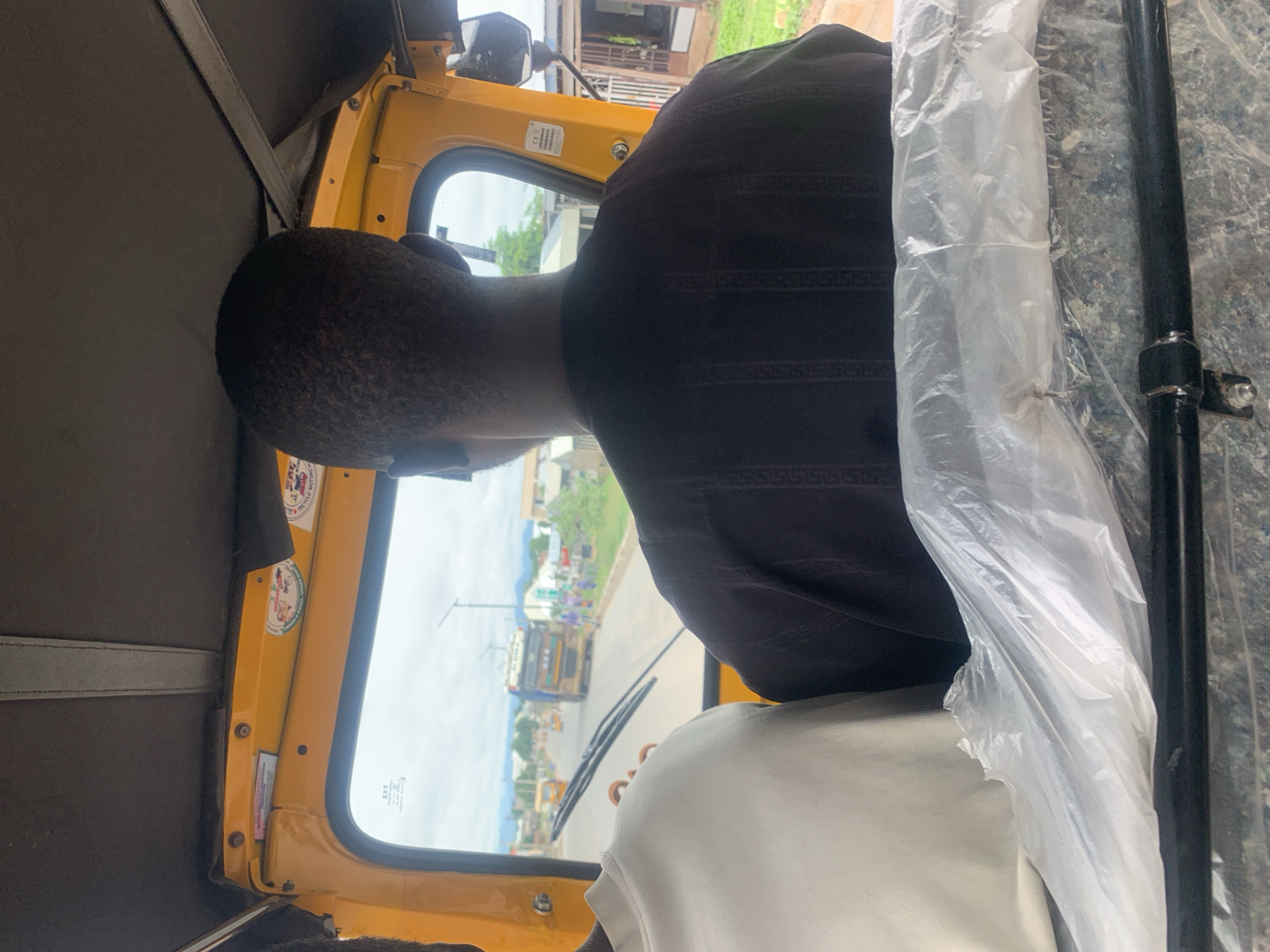 The image shows the interior of a yellow auto-rickshaw in Jalingo, Nigeria. A man wearing a dark patterned shirt is driving, seen from behind. To his left, another person wearing a white shirt is visible. The windshield offers a view of a street with other auto-rickshaws, a large truck, and buildings. The sky is cloudy. There are stickers on the interior of the auto-rickshaw.