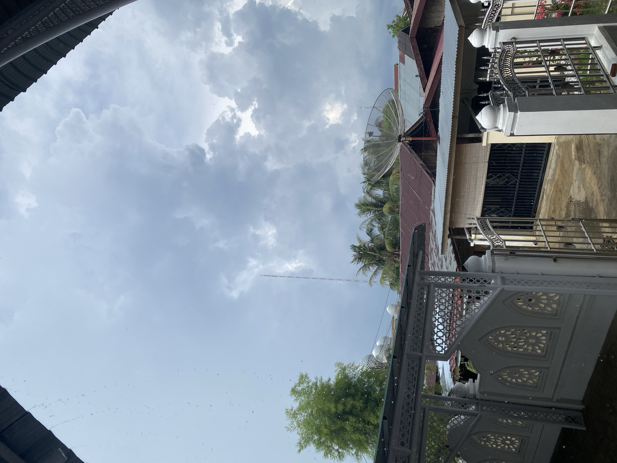 The image presents an upward-looking view of an urban landscape under a heavily overcast sky, with visible rain or recent rain activity. The sky is filled with thick, grey clouds, indicating gloomy or stormy weather. Numerous small water droplets are suspended in the air throughout the frame, suggesting it is either currently raining lightly or has just finished.
In the foreground, a decorative white fence or railing with intricate geometric cutouts and arched patterns is prominently featured. To the right, another section of a building shows a fence with metal railings and white, dome-like finials, hinting at a distinct architectural style.
The midground reveals several buildings with various roof types, including corrugated metal and reddish-brown tiles. A large, white parabolic satellite dish is mounted on one of these roofs. Interspersed among the buildings are lush green palm trees and other foliage, characteristic of a tropical environment.
In the distance, a tall, slender antenna or communication tower rises against the cloudy sky. The architectural details, especially the decorative white structures and patterned railings, combined with the tropical vegetation, suggest a setting in Southeast Asia, aligning with the provided context of Lhokseumawe, Indonesia. The time of day is daytime, but the dense cloud cover creates a diffuse, subdued light. There is no visible text in the image.