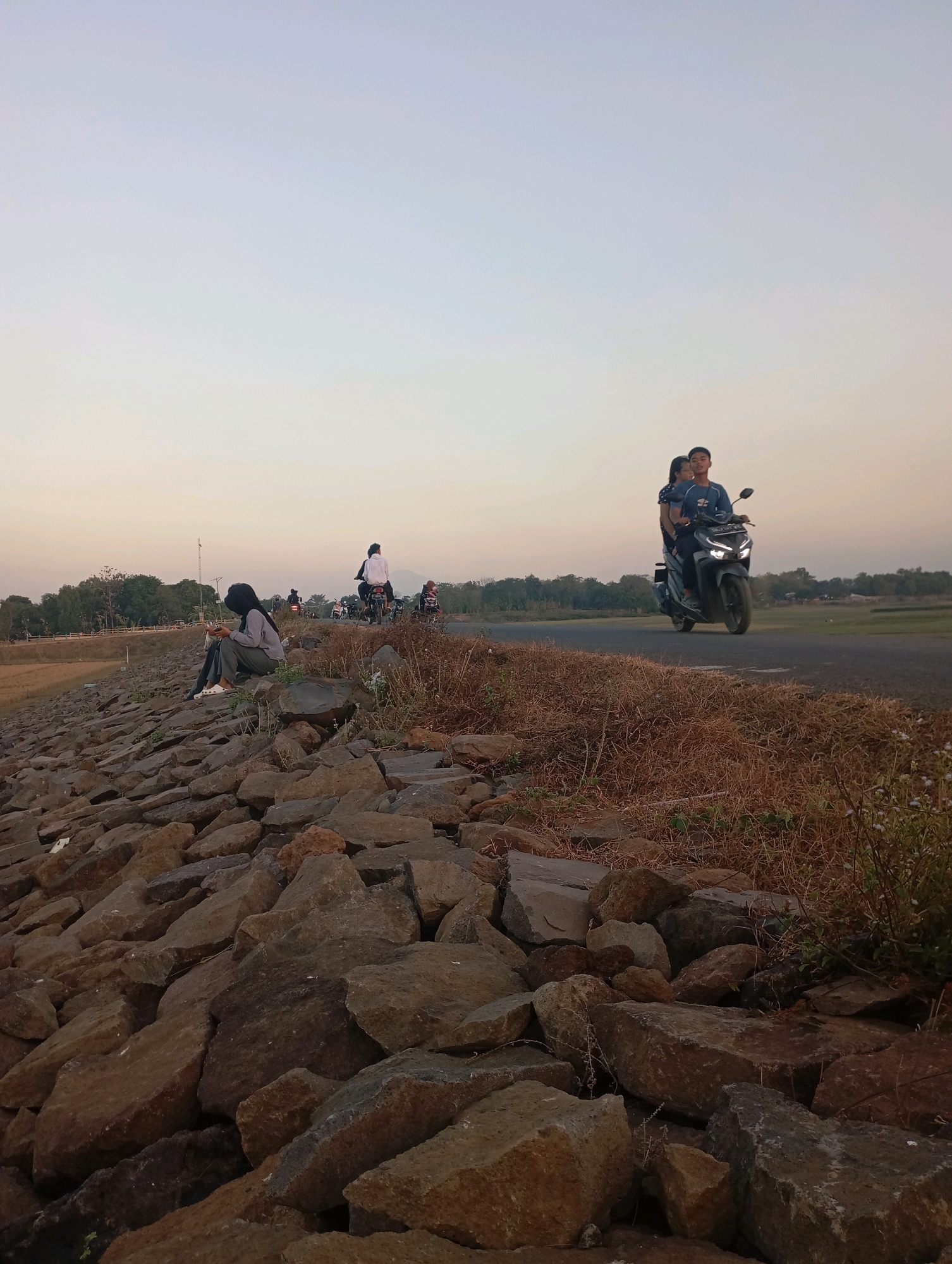An outdoor, low-angle shot captures a dusty road and a rocky embankment in the city of Haurkolot, Indonesia. The sky is a pale gradient of blue and white. On the right side of the road, two people ride a dark-colored scooter. They are moving away from the viewer, with the passenger's arms wrapped around the driver. The driver is a male, wearing a dark blue t-shirt. The passenger is a female, wearing a blue and white polka dot shirt. The road is lined with dry, brown grass and some small flowering plants.
On the left side of the image, a rocky embankment descends towards the viewer. A young person, wearing a gray hoodie and a black hijab, sits on the rocks, looking down at a mobile device. Their legs are crossed, and they are wearing white socks with black slides. Behind this individual, several other people and motorbikes are visible on the road in the distance. Trees and sparse vegetation line the horizon. The rocky embankment is covered in large, irregular stones of varying shades of gray and brown. The ground beyond the embankment appears to be a field of dry, golden grass.