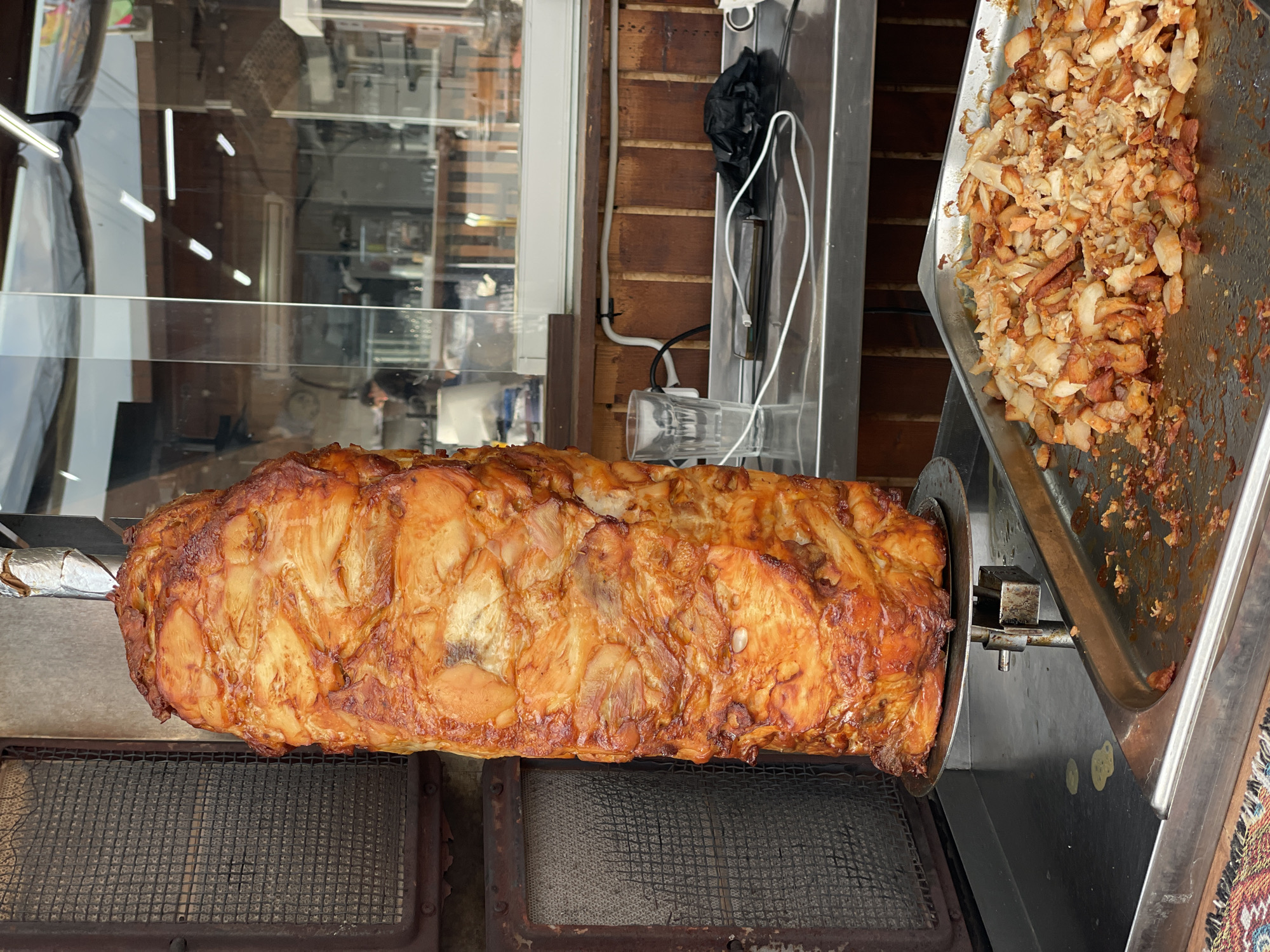 This media file captures a close-up view of a large, rotating spit of seasoned meat, likely chicken, being cooked in an outdoor food stall. The meat is golden brown and appears succulent, with visible layers and some charring from the heat. Below the rotating spit, a metal grate covers what appears to be a heat source. To the right of the meat spit, a stainless steel tray is filled with freshly shaved pieces of cooked meat, ready to be served. The background shows a wooden wall and a glass enclosure, suggesting the stall is set up in a public area, possibly a street or market.
A black plastic bag hangs from a hook on the wooden wall, and electrical wiring is visible, indicating the cooking equipment is powered. The setting seems to be an outdoor establishment, given the natural light and the style of the food preparation. There are no people visible in the frame. The overall impression is one of food preparation in a casual, street-food environment.
The location context provided is Sochi, Russia. There is no visible text in the image. The time of day is likely daytime due to the lighting. The weather is not discernible from the image. The scene evokes a sense of hunger and anticipation for freshly cooked meat.