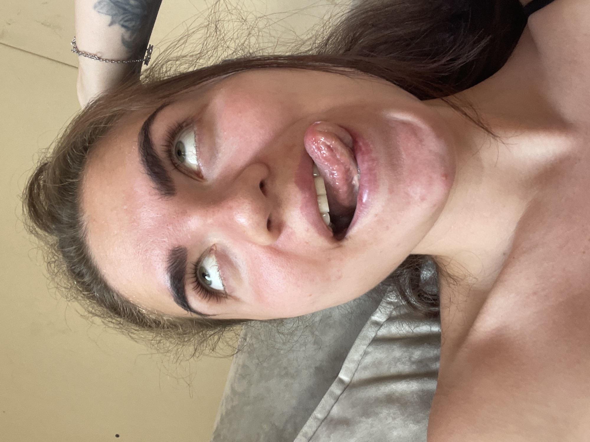 The image is a close-up, slightly overhead shot of a young woman with light brown hair pulled back. She is looking upwards with her eyes wide and her mouth open, her tongue extended and touching her lower lip. Her skin is flushed and appears slightly irritated, with visible pores and some blemishes on her chin. She is wearing a dark bra strap visible on her left shoulder. A silver bracelet with a small rectangular charm is on her right wrist. The background is a plain, light-colored wall and a textured, gray surface that appears to be a cushion or pillow. There is a faint, dark speck on the wall to the left of her head. The image appears to have been taken indoors, likely in a residential setting. There is no visible indication of the location being in Мосметаллоконструкция Онт, Russia.