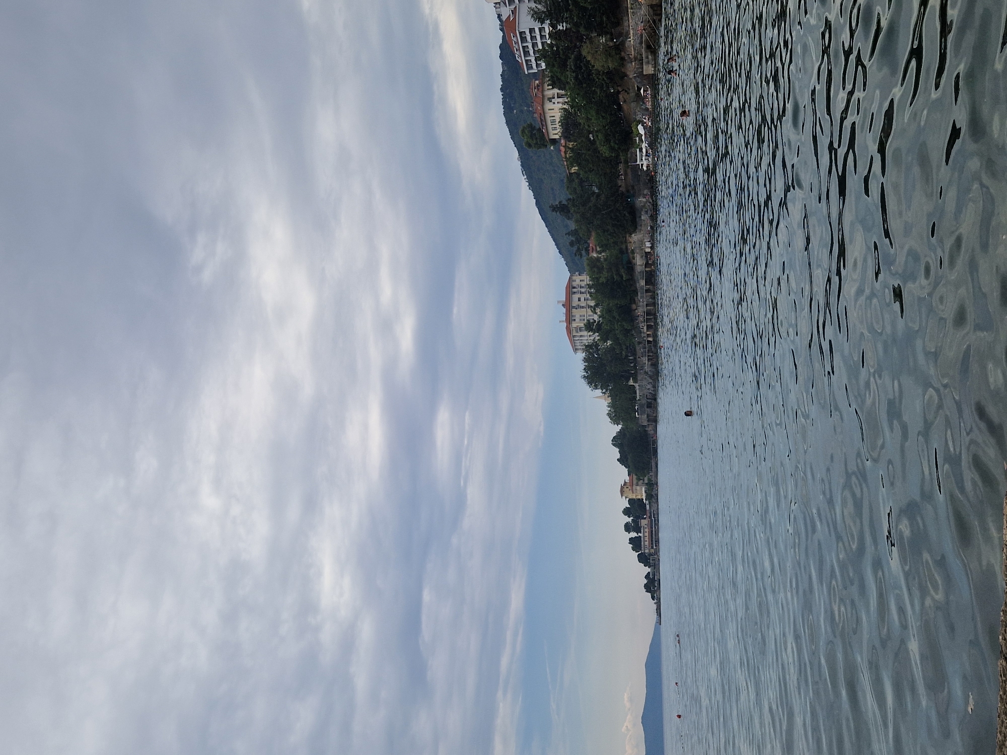 This image depicts the coastline of Ika, Croatia, under a cloudy sky. The foreground is dominated by the choppy, dark blue water of the Adriatic Sea. In the mid-ground, a rocky shoreline is visible, lined with trees and several multi-story buildings, some with red-tiled roofs. These structures appear to be hotels or residential buildings. Further in the distance, a green, tree-covered hill rises behind the buildings. Several people are visible in the water, appearing to swim or relax. Two orange buoys are also present in the sea. The overall scene is a coastal view with natural and man-made elements under an overcast sky.