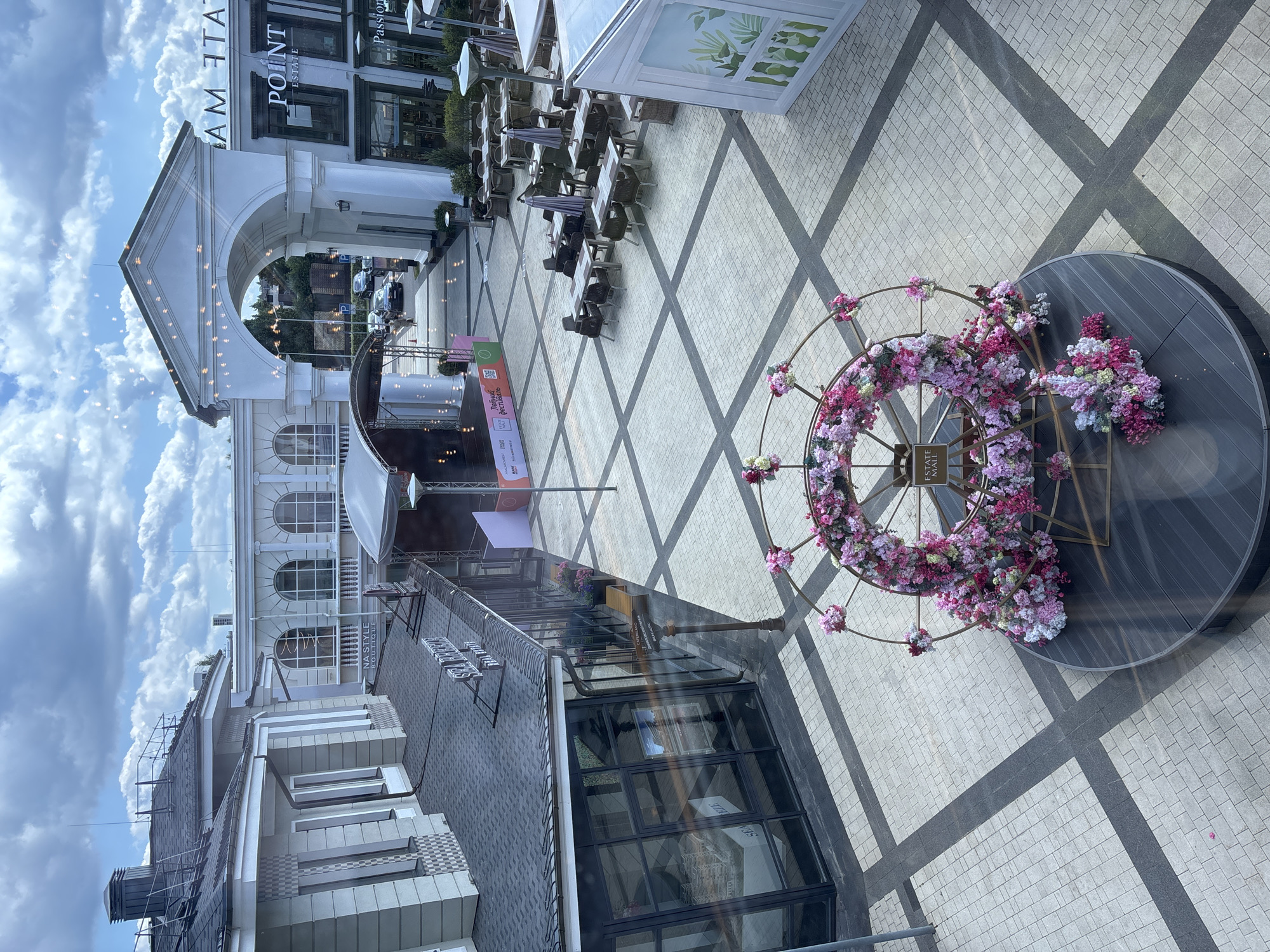 The image captures an outdoor plaza in Monolit, Russia, bathed in daylight under a partly cloudy sky. In the foreground, a large, circular floral decoration, resembling a Ferris wheel adorned with pink and purple flowers, stands prominently on a dark circular platform. The plaza's ground is paved with light-colored square tiles and dark gray square tiles arranged in a diamond pattern.
To the left, a building with a gray facade and dark tiled roof features large glass windows. Above one of the windows, a sign reads "SEA BREEZE." Further to the left, another building section has a lighter facade with decorative columns and arched windows.
In the mid-ground, a stage with a curved roof is set up, displaying various banners and logos, including one with Cyrillic text that appears to say "ЛЮБИМЫЙ ФЕСТИВАЛЬ" (beloved festival). In front of the stage, several tables and chairs are arranged, suggesting an outdoor seating area for cafes or restaurants.
Further back, an arched entranceway dominates the scene. Above the arch, text is visible, with "AM TATE" partially readable on the left and "POINT ESTATE" and "Passion" visible on the right side of the entrance. Cars are seen passing through the archway. The overall environment suggests a well-maintained commercial or entertainment district, possibly a shopping mall or a recreational area.