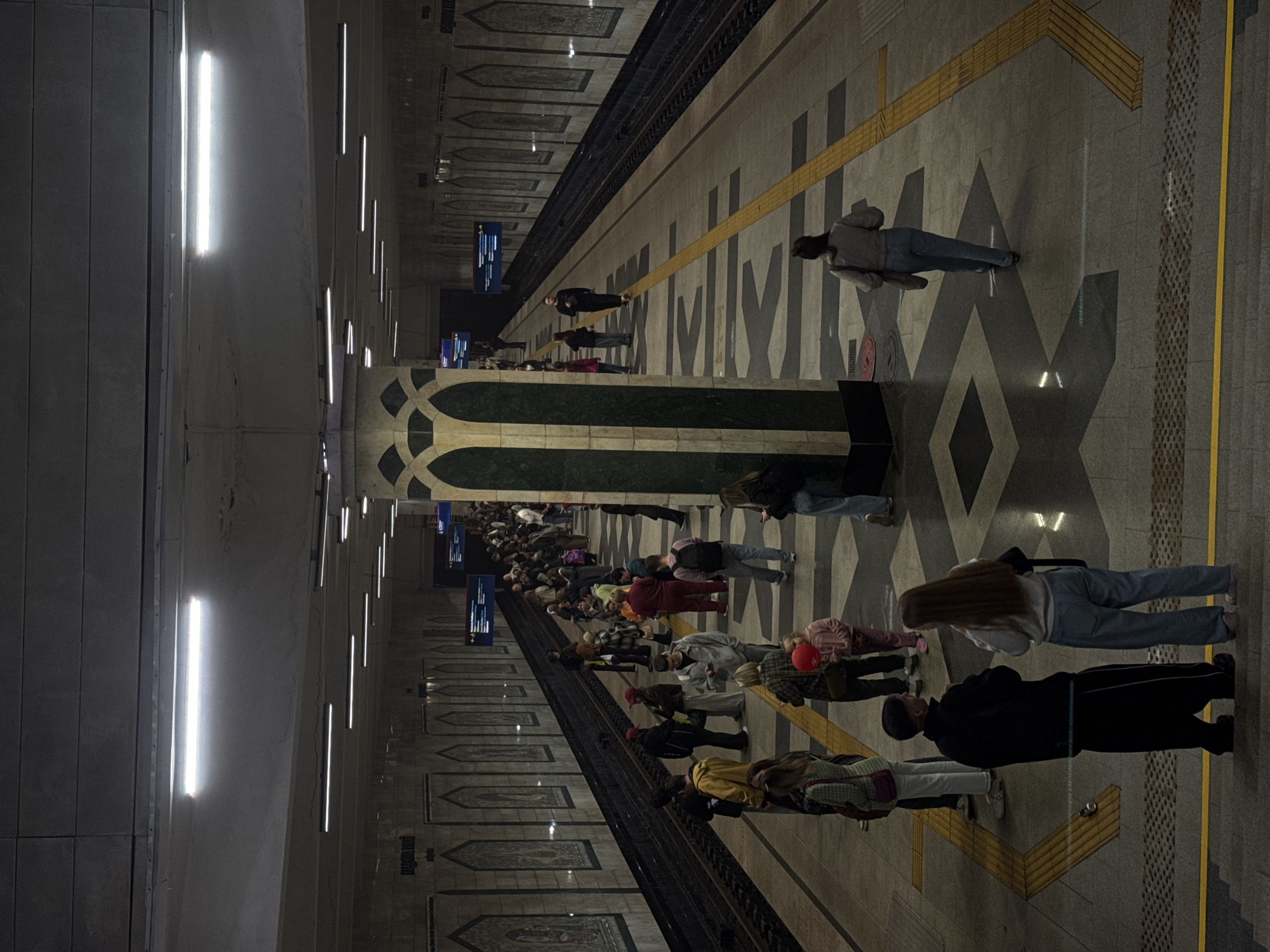 The image shows a crowded subway platform in Kazan, Russia. Many people are waiting along the platform's edge, near a yellow safety line, facing towards the unseen tracks. A few people are standing away from the edge. The platform is tiled with a patterned design of geometric shapes. Along the wall are decorative mosaic panels. Above, a series of bright fluorescent lights illuminate the area. Two electronic display boards are visible in the distance, showing information about train schedules.