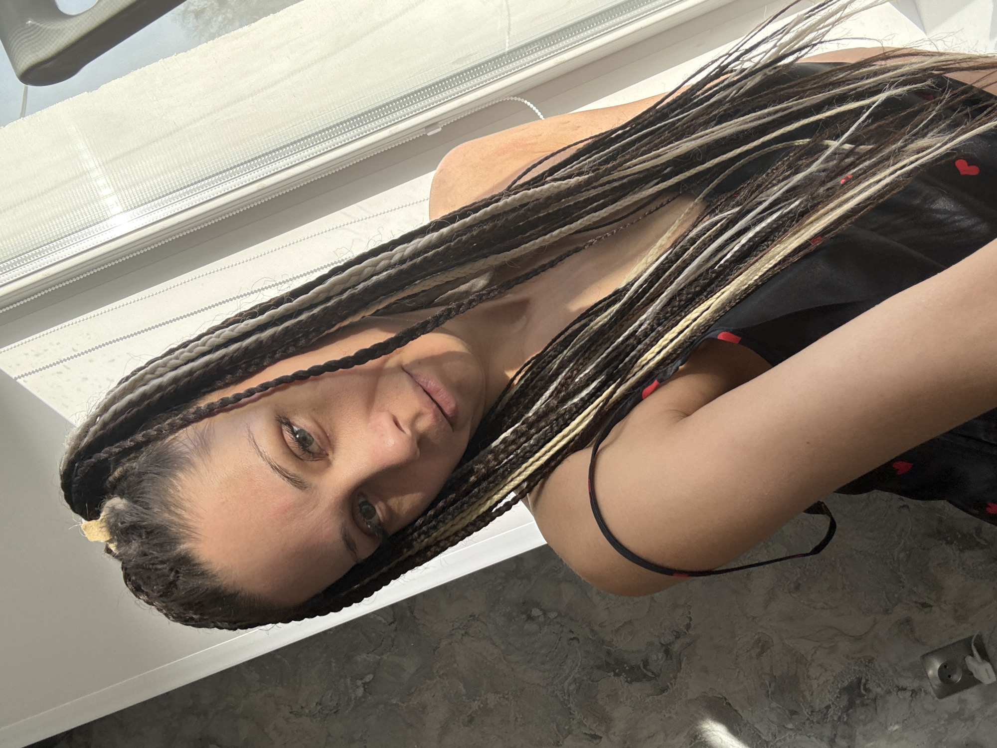 A medium shot depicts a woman indoors, lying down against a white wall. Her head is tilted toward the camera, showing her face in detail. She has fair skin, grey eyes, and long, braided hair with streaks of white and brown. She wears a black garment with small red heart patterns. The background includes a window with a partially visible blind. The setting is inside a building, but specific details about the location, including the city, are not identifiable from the image.