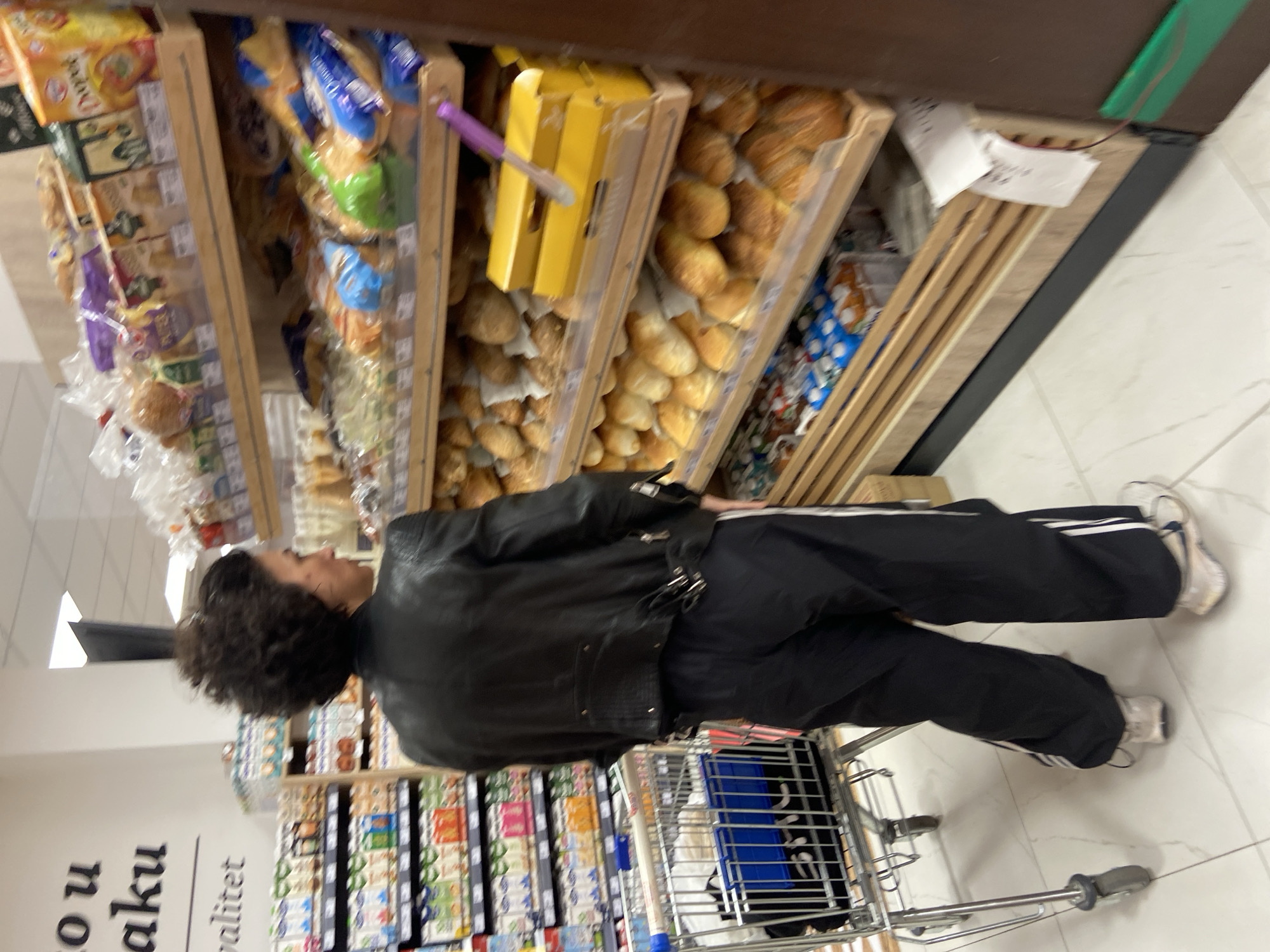 An interior view of a supermarket in Budva, Montenegro, shows a single individual standing in an aisle. The person, an adult of indeterminate gender, is viewed primarily from the back and partial right profile. They have dark, curly, voluminous hair. They are dressed in a black leather jacket with visible zipper and buckle details on the back and sides, black loose-fitting track pants featuring a single white stripe down each outer seam, and light-colored athletic shoes with dark accents.
To the left of the individual is a silver-colored metal shopping cart with a blue plastic child seat, containing indistinct items. To the right, and directly in front of the individual, is a multi-tiered shelving unit displaying a variety of baked goods. The shelves, appearing to be made of light-colored wood, hold numerous loaves of bread, rolls, and packaged pastries in clear plastic. A purple utensil, possibly tongs, rests on a middle shelf. Several yellow boxes are also present on the shelves.
In the background, to the left, another shelving unit is visible, stocked with numerous cartons or bottles of beverages, likely dairy or juice products, identifiable by their colorful labels. Above this shelving unit, partially visible text on a white wall reads "o u aku" and "valitet," indicating Montenegrin text for "in packaging" and "quality." The floor is covered in white square tiles with dark grout lines. The scene is brightly illuminated by overhead artificial lighting. The individual appears to be examining products in the bakery section.