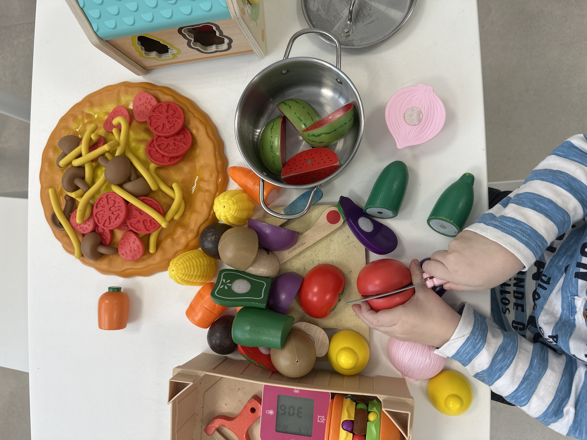 A child is playing with toy food on a white table. The child, wearing a blue and white striped shirt, is using a toy knife to cut a red toy tomato. Around the tomato are other toy fruits and vegetables, including corn, carrots, mushrooms, eggplant, and a pink onion. There's also a toy pizza, a pot with toy watermelon, and a wooden box. The location is unspecified, and there's no evidence the image was taken in Votkinsk, Russia.