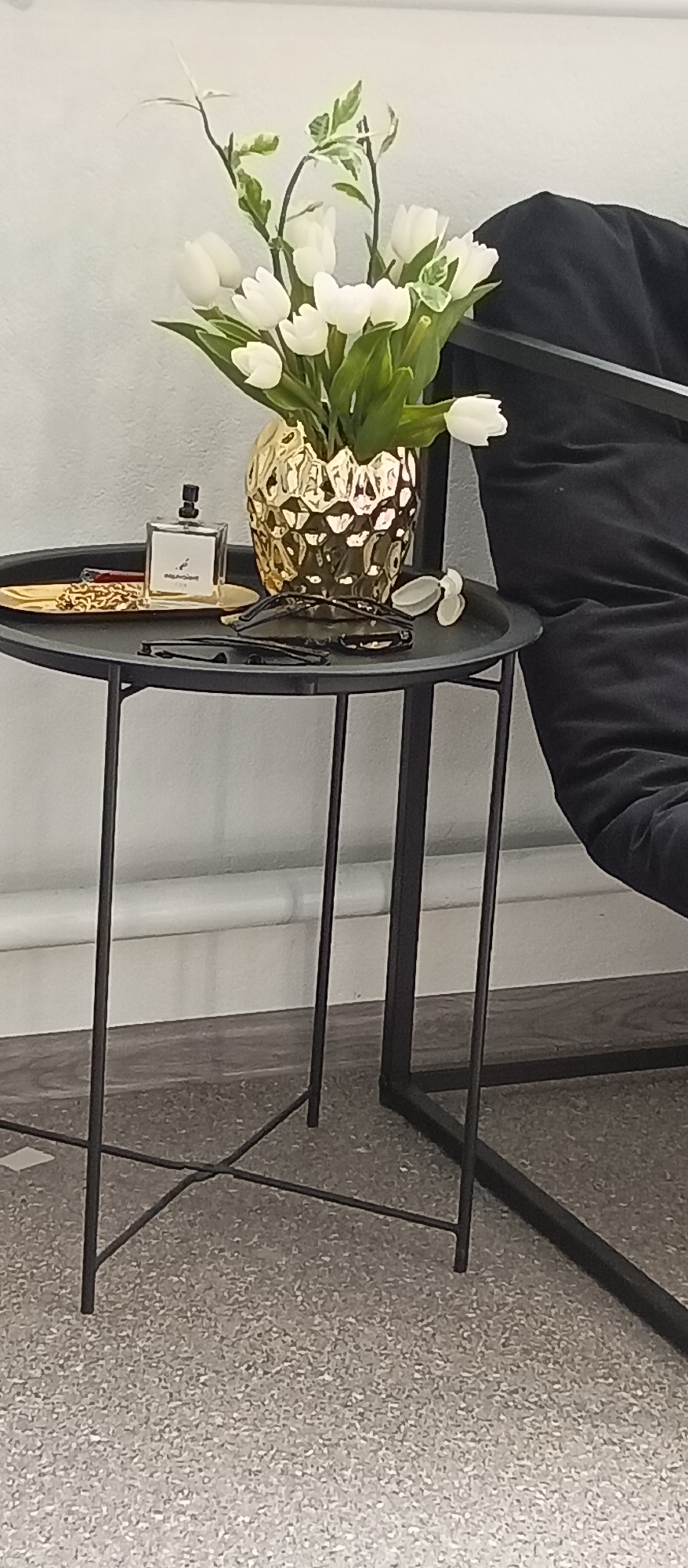A side table, next to a black upholstered chair, holds a gold vase filled with white tulips, a bottle of perfume, a golden tray with jewelry, and eyeglasses. The setting appears to be indoors.