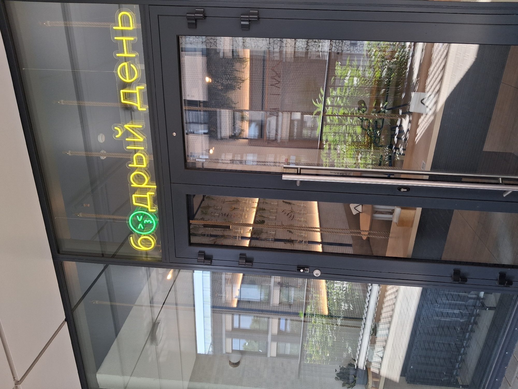 A storefront with a neon sign reading "Бодрый день" (Bodryi den', meaning "Cheerful day") in Cyrillic characters, with a smiling emoji character integrated into the text. The sign is in yellow and green neon. The storefront features large glass panels and dark gray doors with silver handles. Inside, there are hanging plants, wooden furniture, and warm lighting. The exterior shows sunlight and reflections of buildings in the glass. The scene appears to be a cafe or restaurant. The location is in Novosibirsk, Russia.