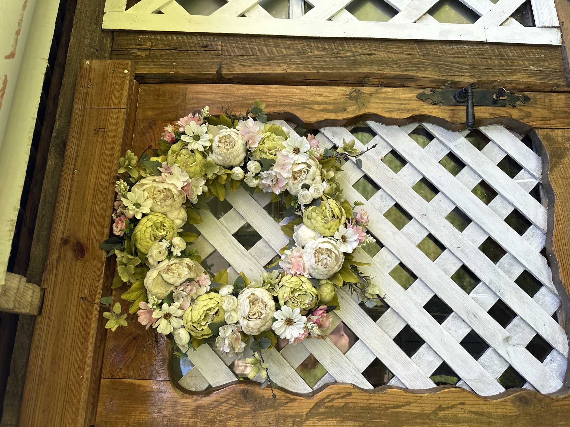 A floral wreath adorns the wooden door of the Norviliškės Manor in Lithuania. The wreath, composed of a variety of faux flowers in shades of cream, pale green, and pink, is hung on the upper portion of the door, which features a decorative lattice insert. The door itself is made of aged, natural wood with visible grain and a patina. To the right of the door is a white lattice structure. The scene appears to be an exterior shot, possibly an entryway.