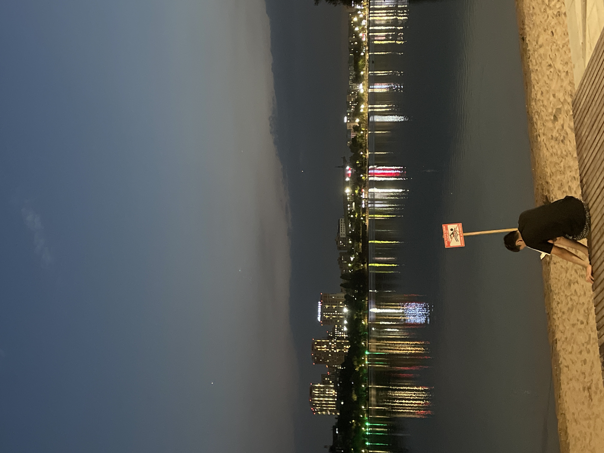 The image depicts a tranquil twilight scene by a body of water in Almaty, Kazakhstan, likely at Kok-Tobe Lake, given the city skyline. In the foreground, a person, viewed from behind, sits on a wooden deck on a sandy shore. The individual is wearing a black t-shirt and patterned shorts and is looking down, seemingly engaged with a handheld device. To the left of the person is a red and white sign on a pole, indicating "No Swimming."
Across the water, the Almaty skyline is illuminated with lights from various buildings, reflecting vibrantly on the calm surface of the lake. The sky is a deep twilight blue, with a few faint stars visible. The overall atmosphere is one of quietude and evening relaxation.