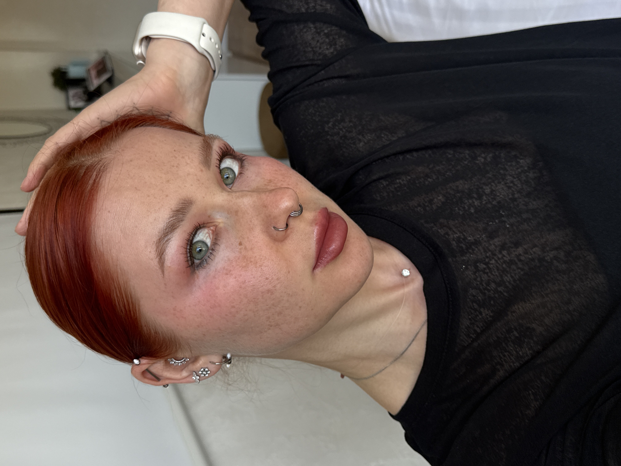A close-up portrait shows a woman with red hair, green eyes, and numerous freckles. She has a white watch on her wrist and her hand is near her head. She has several piercings on her nose and ears. She is wearing a black, long-sleeved top. The background is indistinct, but there is a hint of a room setting with white surfaces. There is no indication of the location being Rostov-on-Don, Russia.