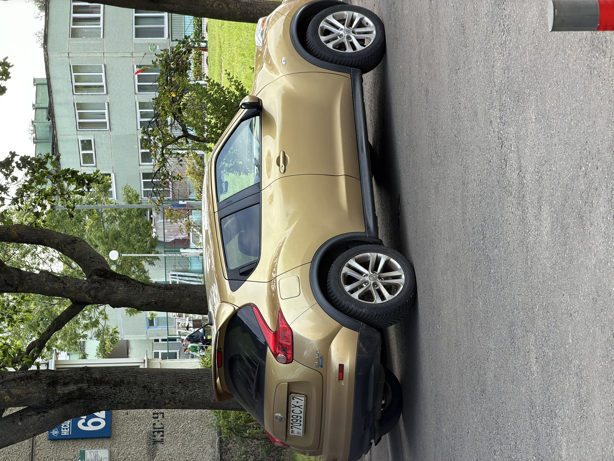 A gold Nissan Juke is parked on an asphalt road in front of a green, multi-story building with numerous windows. The building appears to be an apartment complex or an institutional building, possibly a school, given the presence of a playground visible in the background. Adjacent to the building is a large tree with green foliage, partially obscuring the structure. Another tree, with a rough, dark bark, stands to the left of the car. On the building's facade, a blue sign with white text reads "HЕСЛ" and the number "62". Further to the left, a black inscription on a textured wall reads "КЭЭ-9". Two individuals, a man and a woman, are seated on a bench near the base of the tree on the left. The man is wearing a dark jacket and a hat, and the woman is wearing a green jacket. The license plate on the back of the car is BY 7099 CX-7. The overall environment suggests an urban setting in Minsk, Belarus, during the daytime.