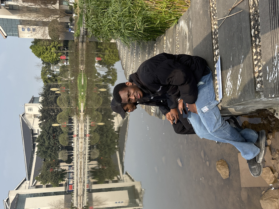 Two young men sit side-by-side near a body of water, likely a lake or pond. The man closest to the camera wears a black puffer jacket and light blue jeans. The other man wears a black jacket and a dark baseball cap. They are positioned on a concrete ledge with stone accents, overlooking the water. Buildings with traditional roofs and greenery reflect in the calm water. The photo is taken during the daytime with natural light.