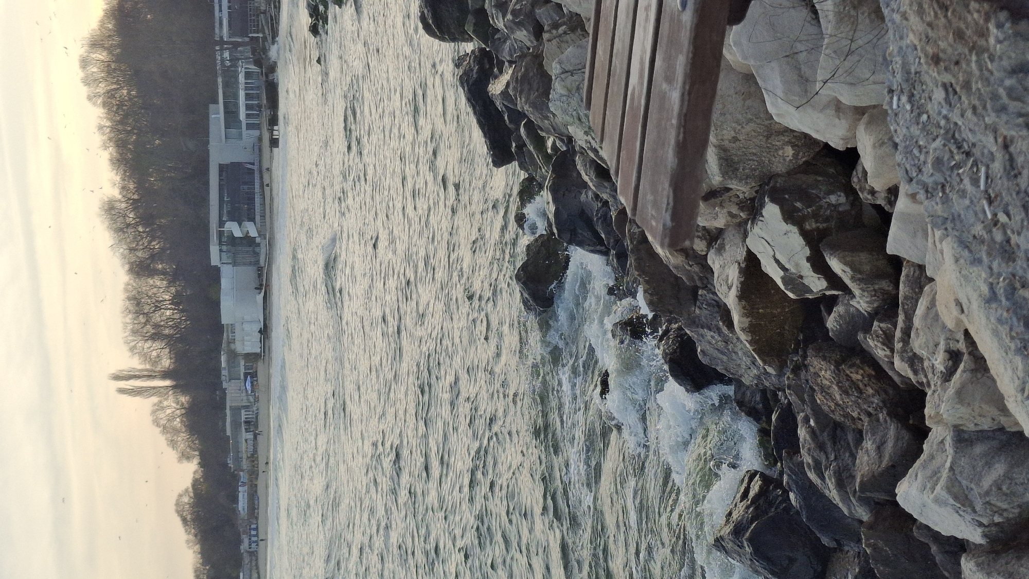 The image depicts a coastal scene with water on the left, a rocky shoreline on the right, and a view of buildings and trees in the background. The water is active with ripples and splashes against the rocks. On the right, there's a section of a wooden structure, possibly part of a bench or walkway, adjacent to the rocks. The background reveals buildings along the coastline, with trees further inland. There are no people visible in the image. Based on the buildings' architecture and general atmosphere, the location is potentially Varna, Bulgaria, considering the area's known coastal features.