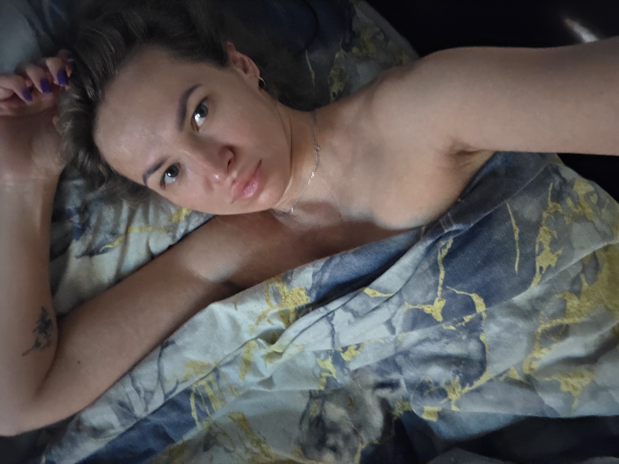 A fair-skinned woman lies on a bed in an indoor setting. She is partially covered by a blue and gold patterned bedsheet. She looks directly at the camera with a neutral expression. Her left arm is raised with fingers near her head. A small tree tattoo is visible on her left forearm. She wears a thin necklace. Location: Possibly Chaikovsky, Russia.