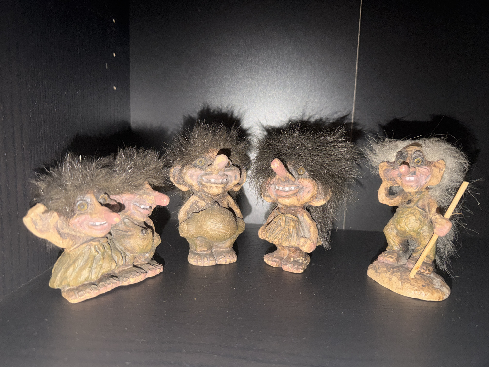 The image features a static display of four distinct troll figurines arranged on a dark, flat surface, likely a shelf, against a deep black background. Bright, frontal lighting illuminates the figures, casting noticeable shadows behind them.
All four trolls exhibit exaggerated features, including large noses, wide, toothy grins, and prominent eyes. Their bodies are textured, appearing to be crafted from ceramic or resin in earthy tones of brown, beige, and muted green. Each troll possesses a mop of shaggy, synthetic hair, varying in color and texture.
From left to right, the first figurine shows two smaller trolls seemingly intertwined or closely grouped, both having dark, spiky hair and cheerful expressions. The second troll is a singular figure with dark, shaggy hair, a pronounced potbelly, and a broad smile. The third troll also stands alone, characterized by dark hair and a similar grinning face, appearing to wear a skirt-like garment. The fourth and final troll on the right has lighter, greyish hair and a more impish expression, clutching a slender, light-colored staff or stick in its right hand.
The setting is an indoor space, possibly a home or shop in Sofia, Bulgaria, as indicated by the provided context. There is no visible text or ongoing activity, emphasizing the decorative nature of the display.