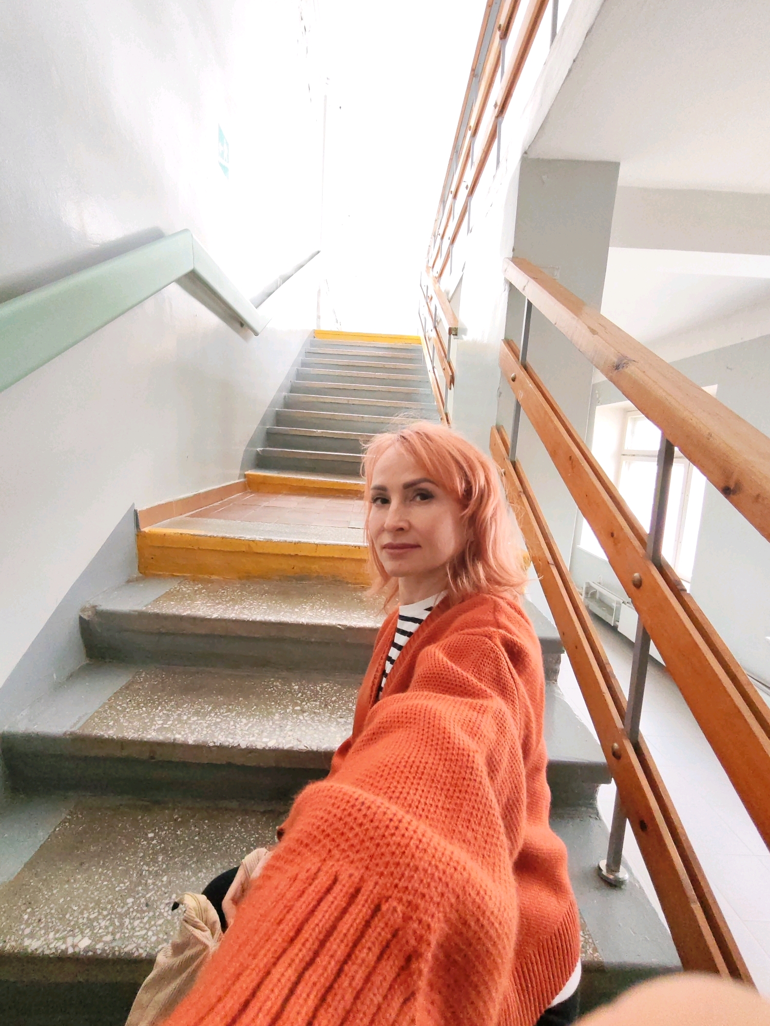 A woman with peach-colored hair, wearing an orange knitted cardigan over a striped shirt, takes a selfie on a concrete staircase with yellow-painted risers. She is positioned on the stairs, looking at the camera. The staircase has wooden handrails. The environment appears to be an indoor stairway in a building, possibly residential or an office building, located in the city of Chaikovsky, Russia. The lighting is bright, suggesting daylight coming from above.