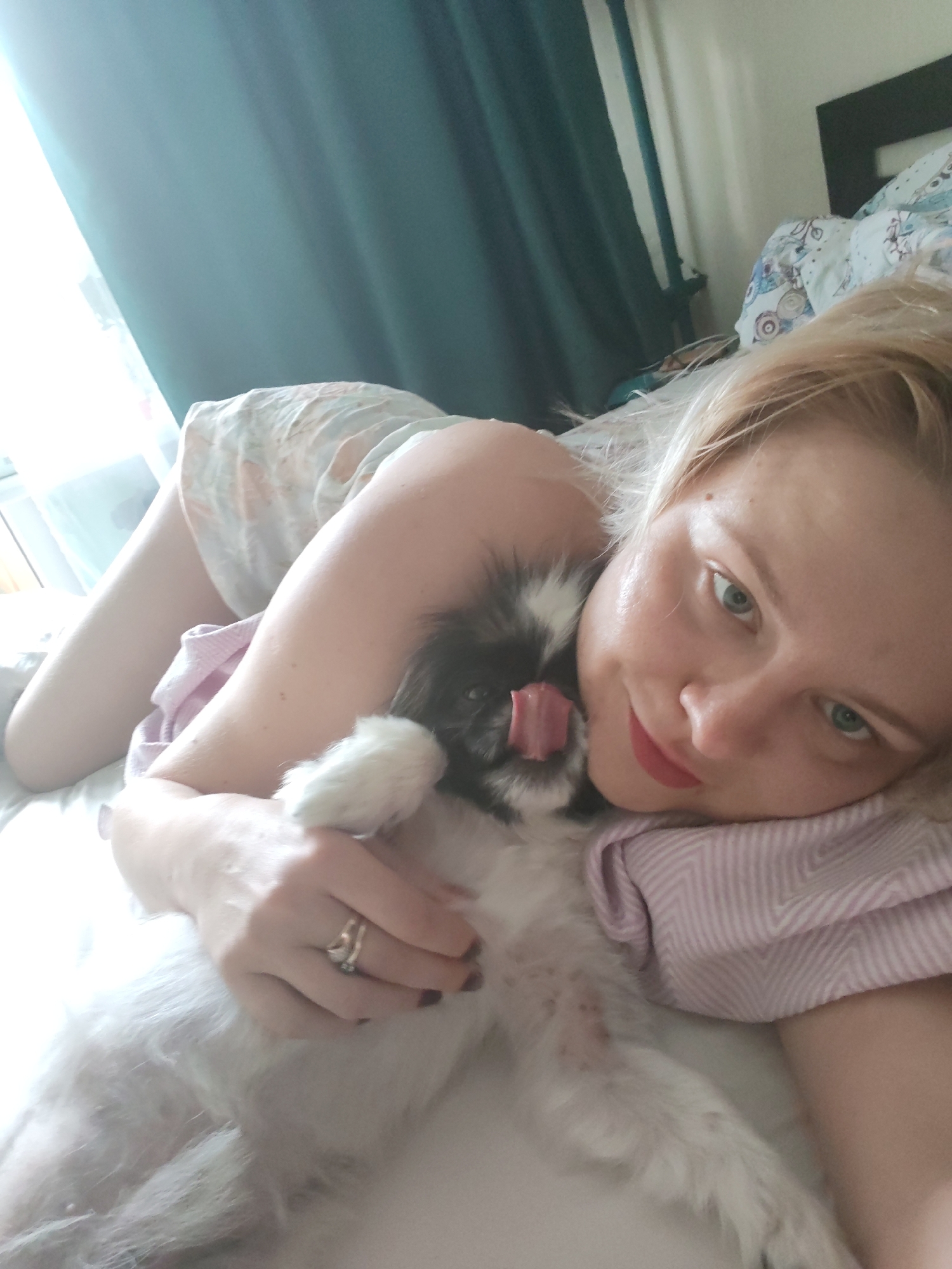 A person with blonde hair is lying on a bed with a dog. The person is on their side, facing the camera, with their head resting on a pillow. The dog is black and white, with its tongue sticking out, and is lying on top of the person. The person is wearing a light-colored top with a floral pattern. The environment appears to be a bedroom, with a window and curtains visible in the background. The bed has patterned sheets and pillows. The image was taken in Izhevsk, Russia.